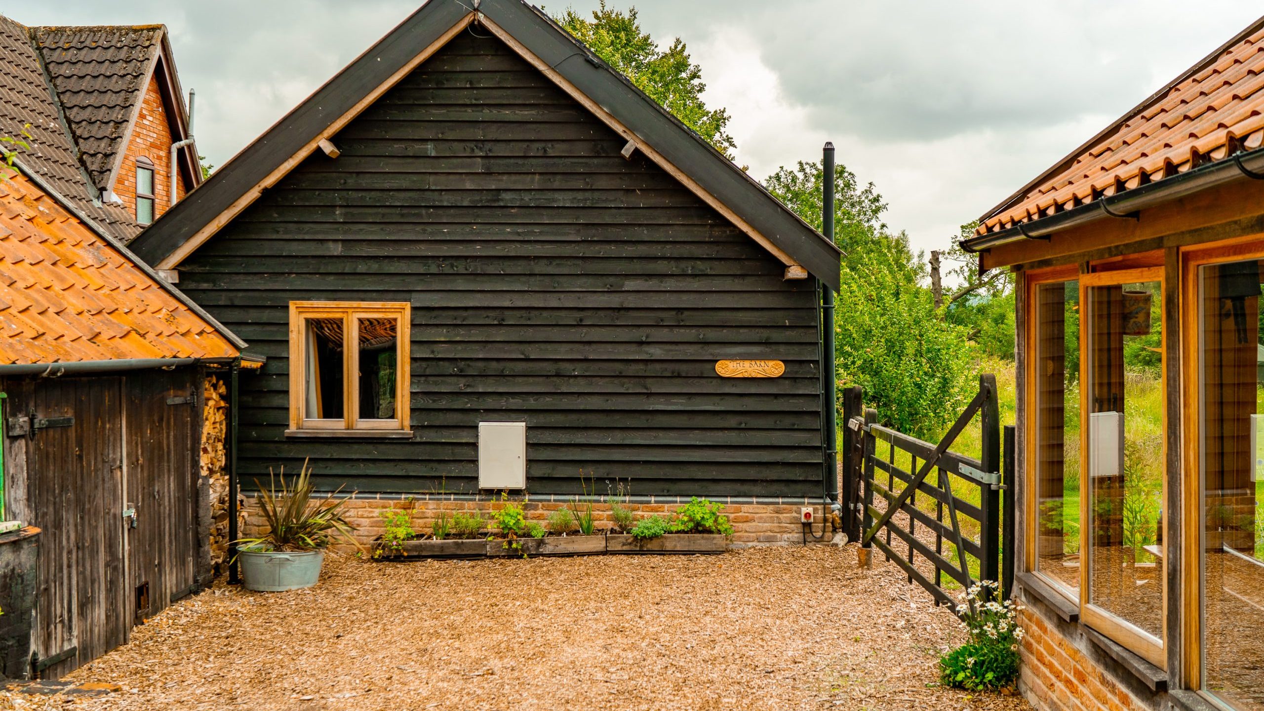 A rustic backyard scene featuring a small, black wooden house with a pitched roof and a single window. The surrounding buildings have red tile roofs. A wooden gate and a garden with small plants are in the foreground. The sky is overcast.