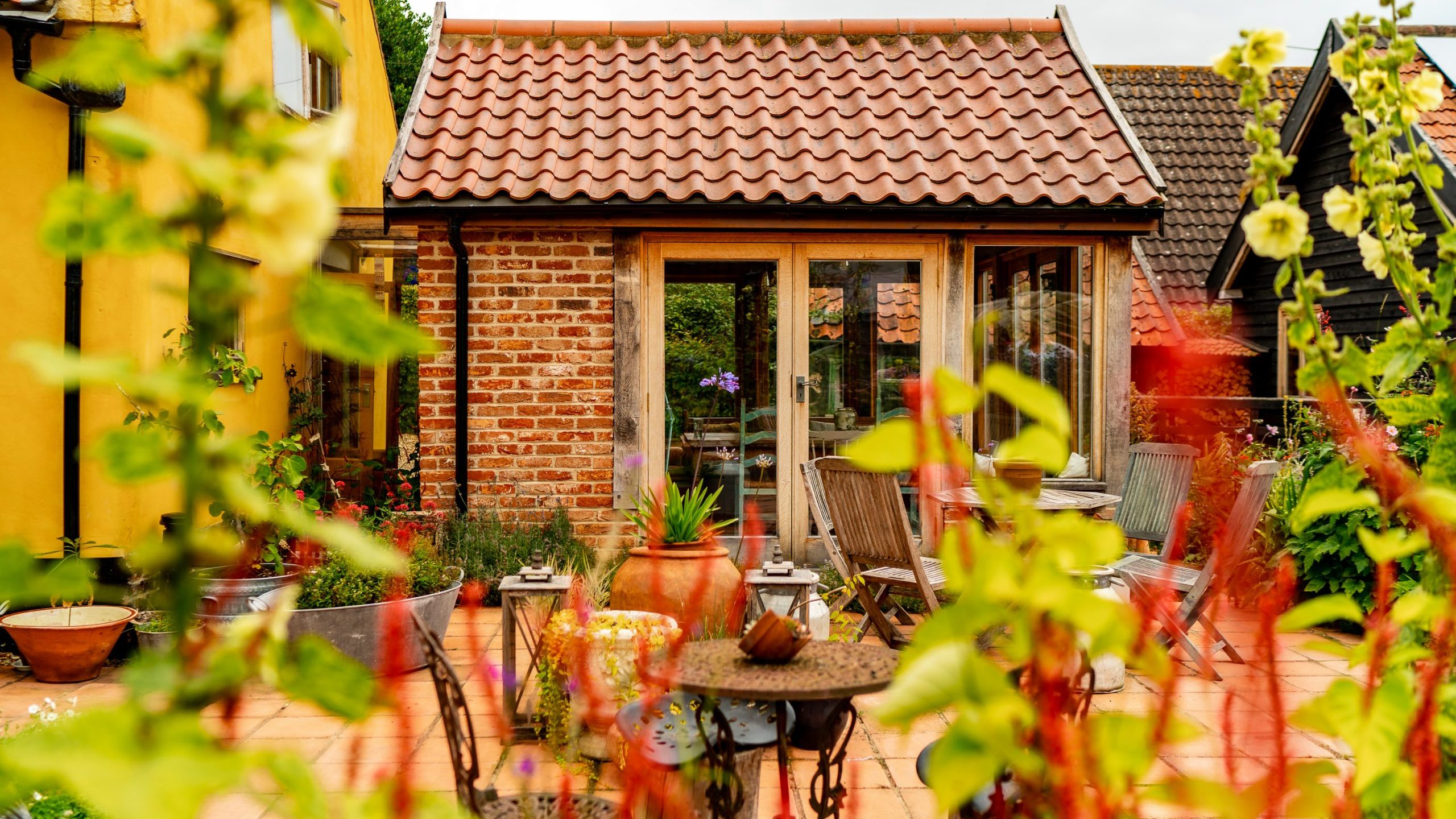 A tranquil backyard scene with a cozy brick and wood house featuring a red-tiled roof. The patio, adorned with various plants and vibrant flowers, has several wooden chairs and a table. The scene is framed by green and red foliage in the foreground.