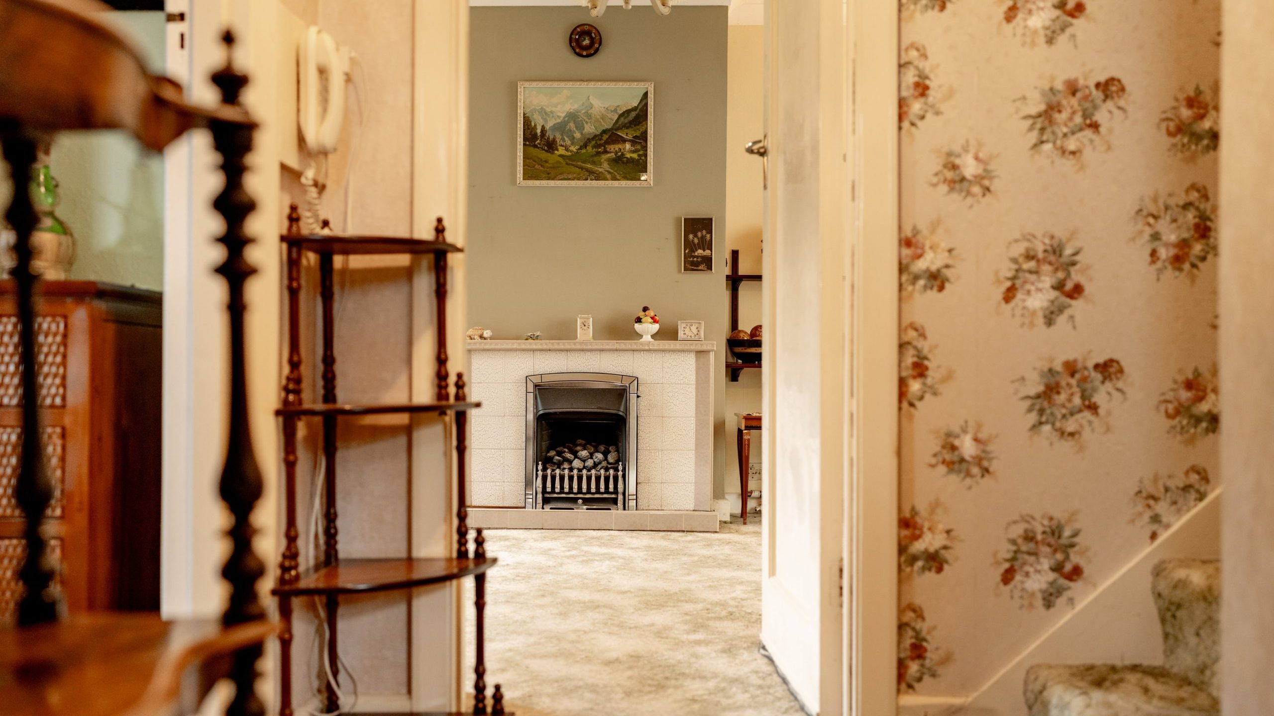 A cozy, vintage-style hallway leads into a living room. The walls feature floral wallpaper, and steps with a carpeted staircase are visible. The living room has a fireplace, a painting of a landscape above it, and a shelf with decorative items and a red teapot.