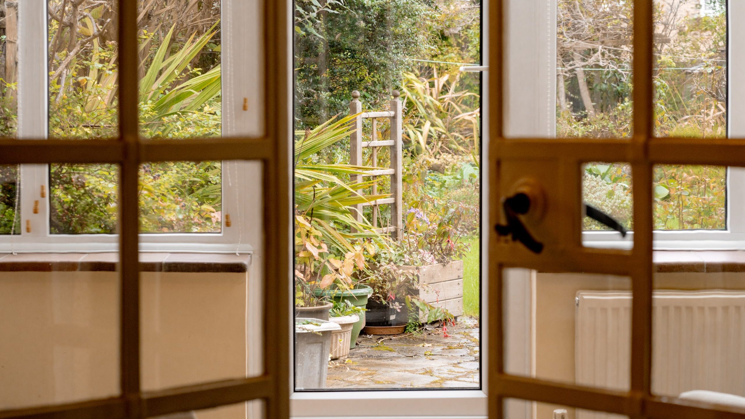 A view through glass doors into a garden, featuring lush greenery and a small path leading outside. The doors have a traditional wooden frame with sections of glass panes, and the garden is filled with plants, shrubs, and a glimpse of outdoor furniture.