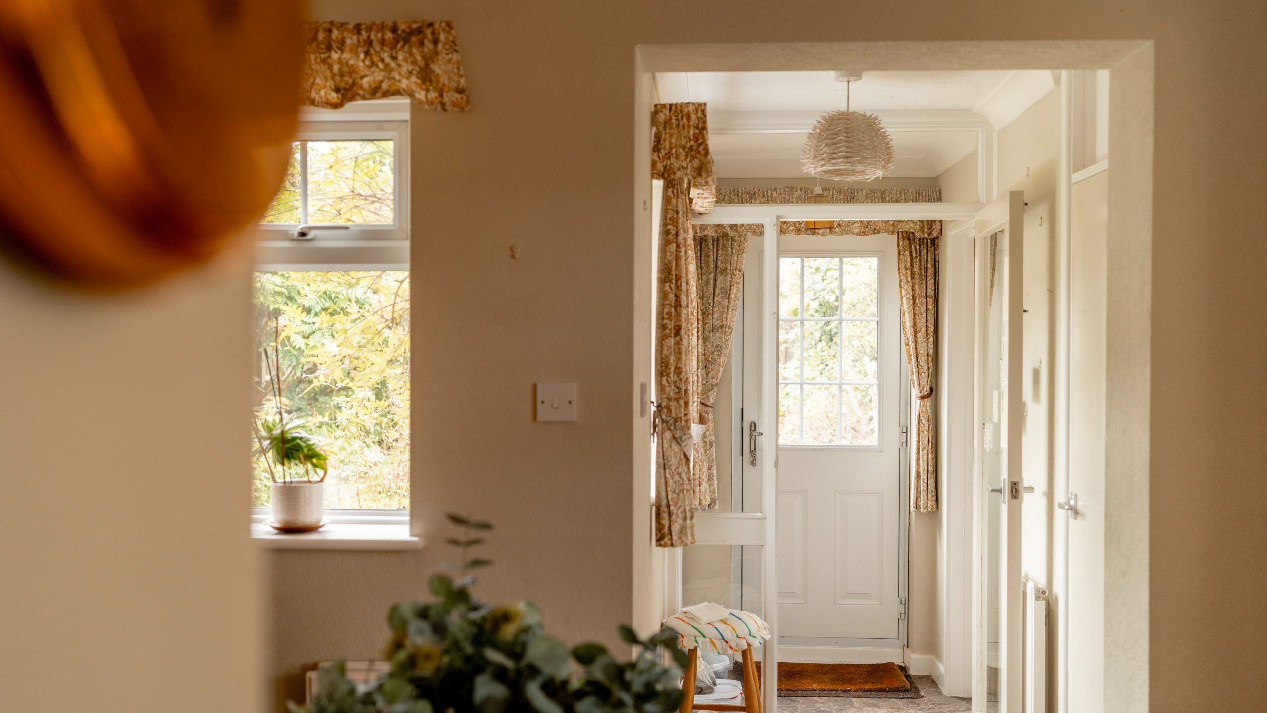 A cozy hallway entrance of a home with natural sunlight coming through a window on the left, illuminating a potted plant on the windowsill. The entry door at the end has a decorative curtain, and there are light-colored walls with a few framed pictures and floral curtains.