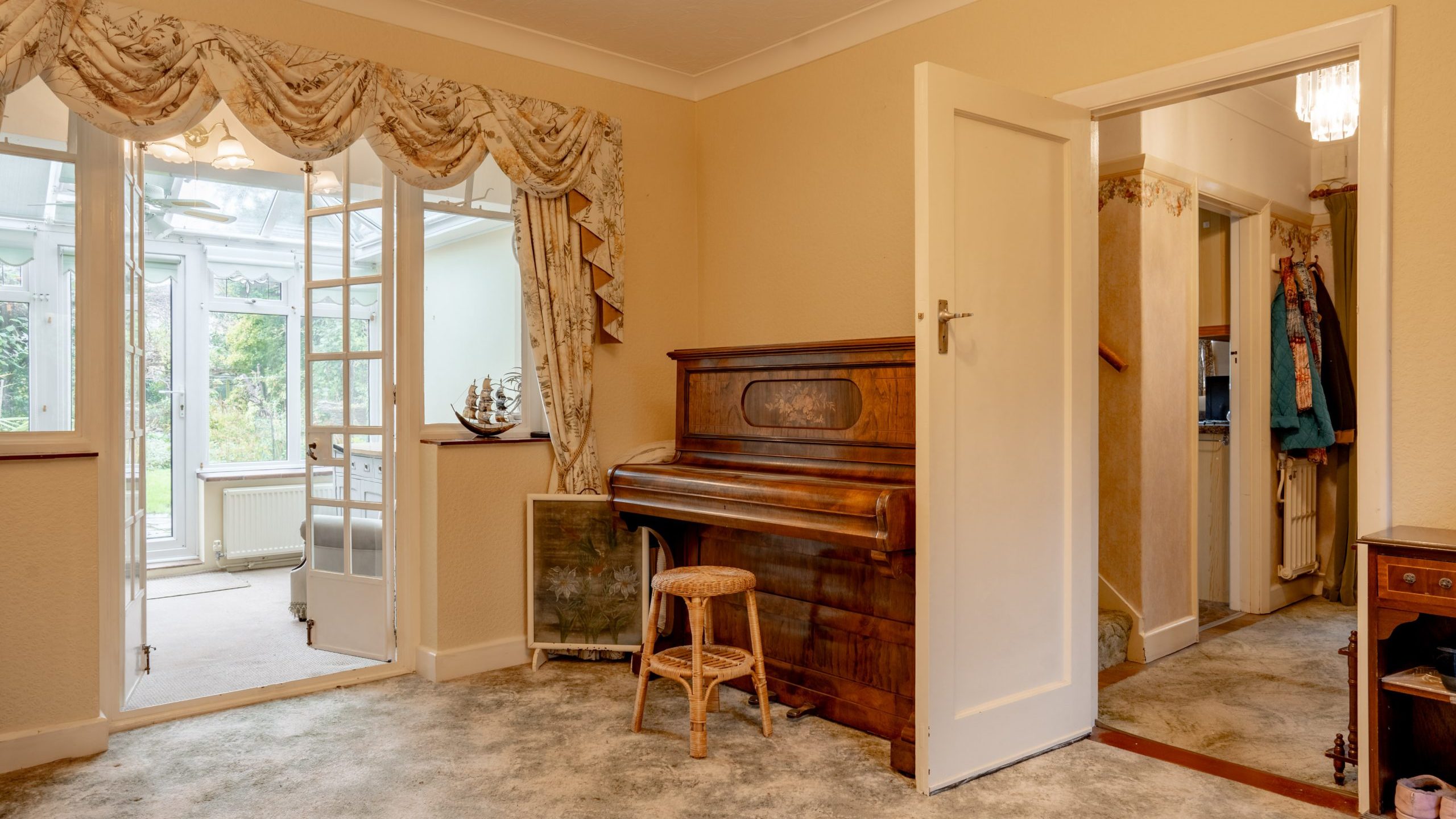 A cozy room with a wooden piano and a wicker stool in front of it. French doors with ornate curtains lead to a bright conservatory with a garden view. An open door reveals a hallway. The room has a light carpet and is decorated in warm tones.