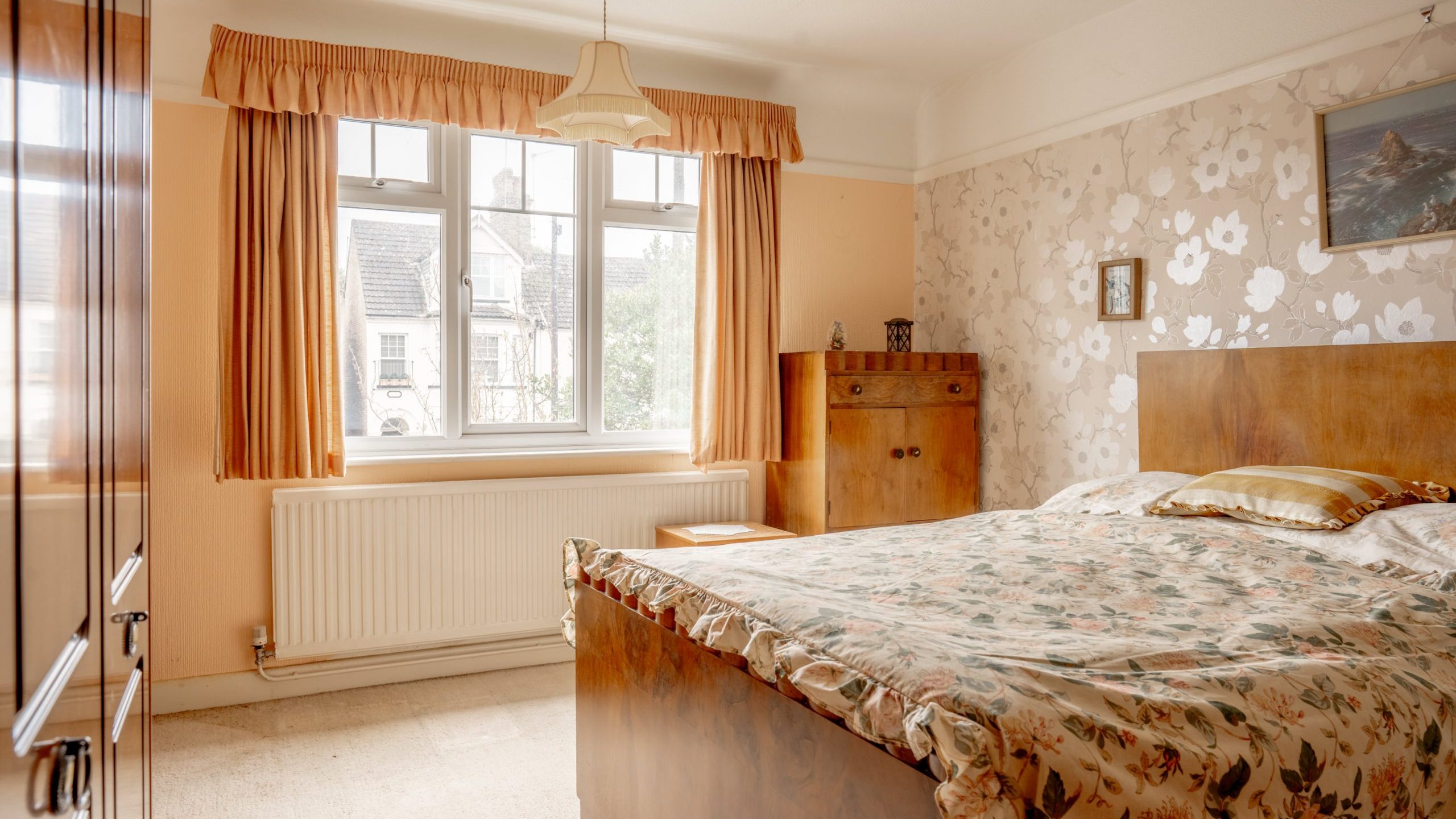 A bedroom with floral wallpaper, peach-colored walls, and a large window with peach curtains. The bed has a floral bedspread, a wooden headboard, and matching wooden furniture, including a wardrobe and a dresser. A ceiling light fixture hangs above the bed.