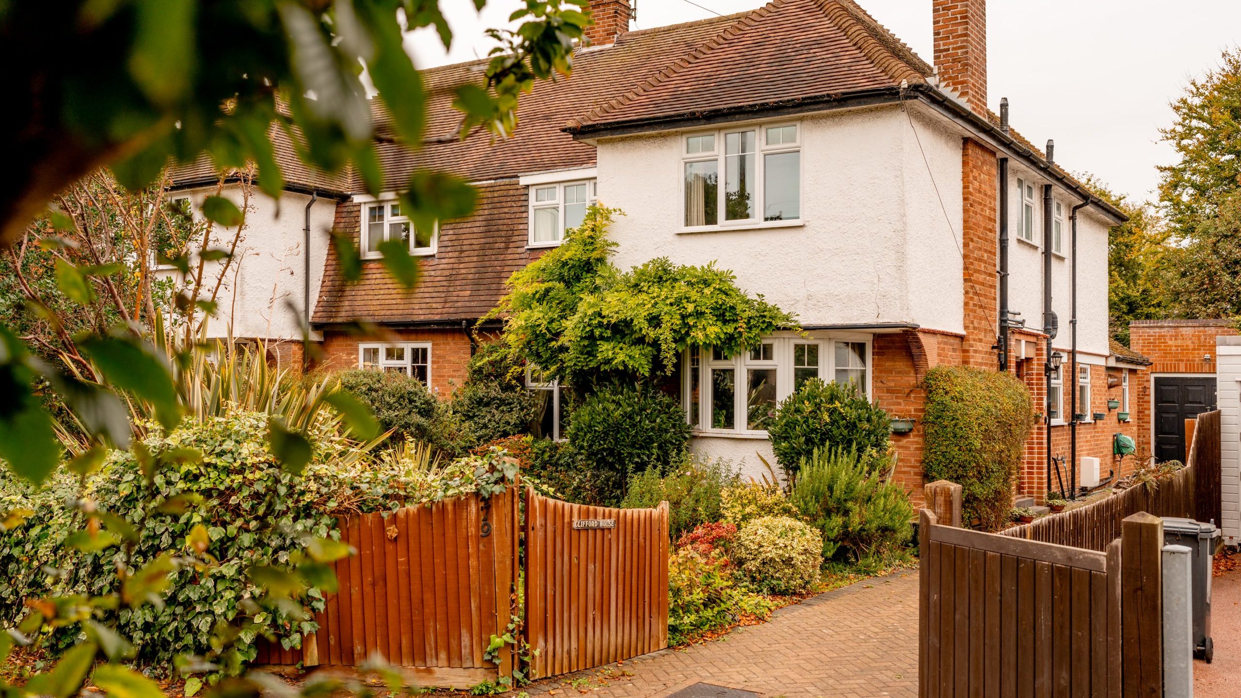 A two-story house with white walls, brown roof tiles, and a brick chimney is partially obscured by greenery. The front yard is filled with various plants and shrubs, and a wooden fence surrounds the property. A narrow brick pathway leads to the front door.