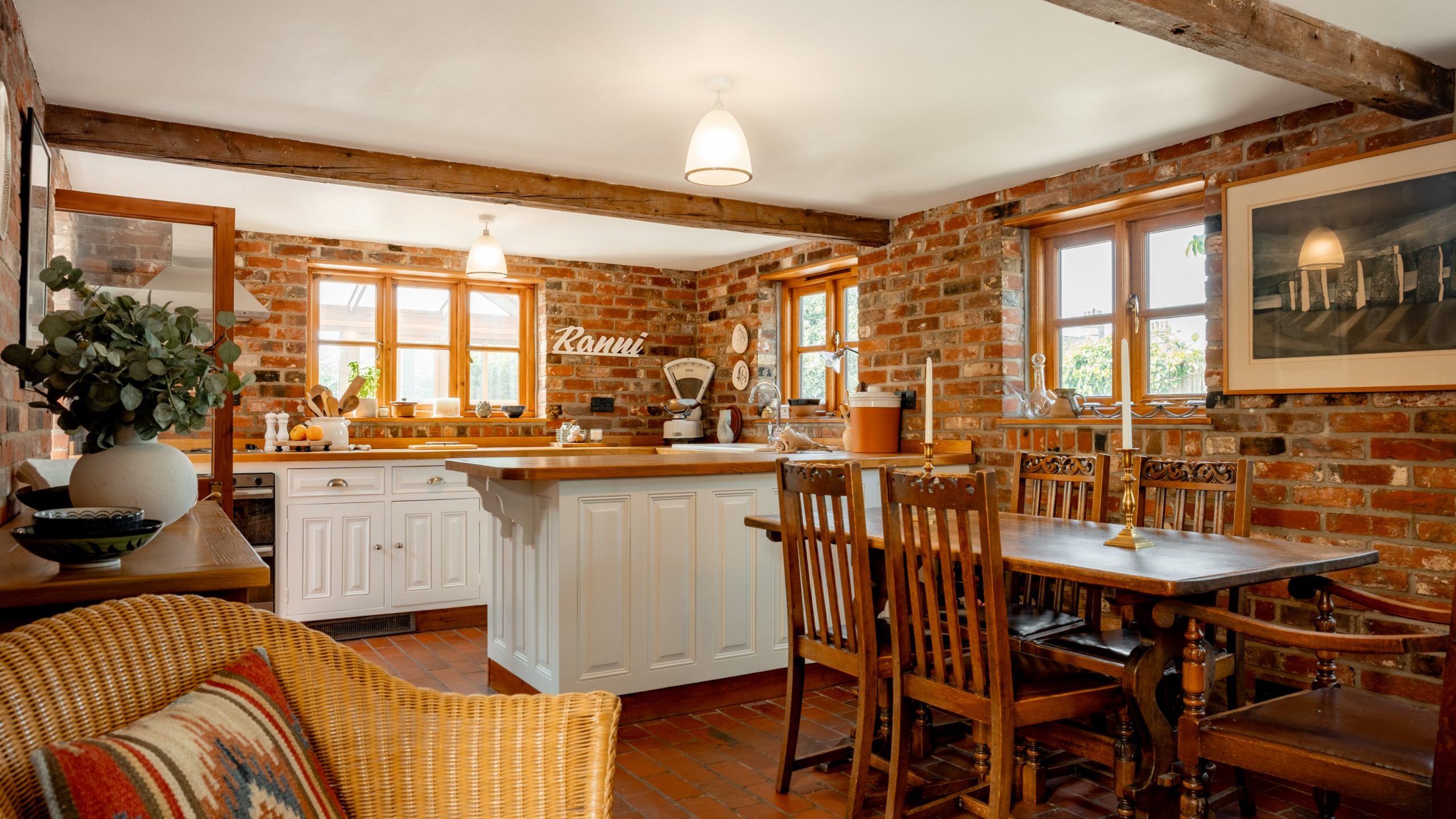 A cozy kitchen with an exposed brick interior features wooden ceiling beams, a white island with a marble countertop, and a rustic wooden dining table with chairs. A wicker chair with a colorful cushion sits nearby, and a large window lets in natural light.