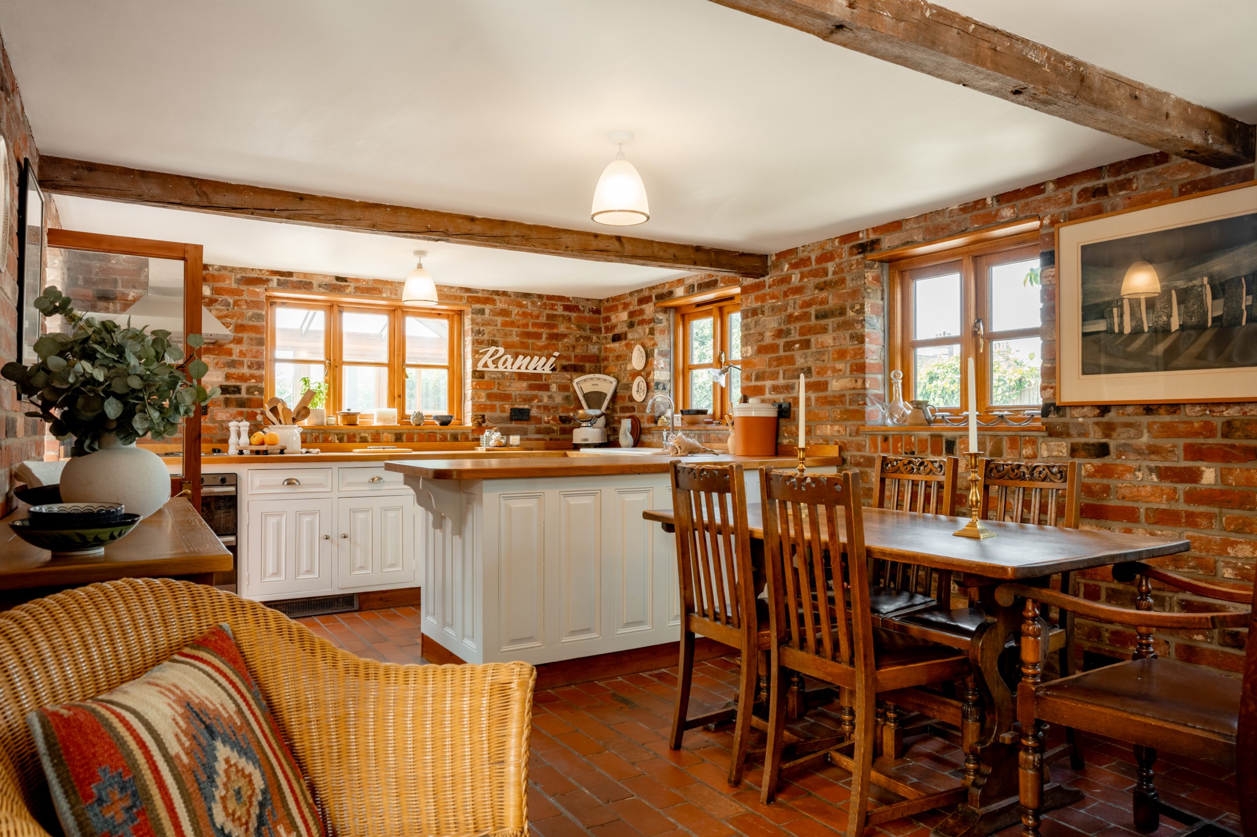 A cozy kitchen with an exposed brick interior features wooden ceiling beams, a white island with a marble countertop, and a rustic wooden dining table with chairs. A wicker chair with a colorful cushion sits nearby, and a large window lets in natural light.
