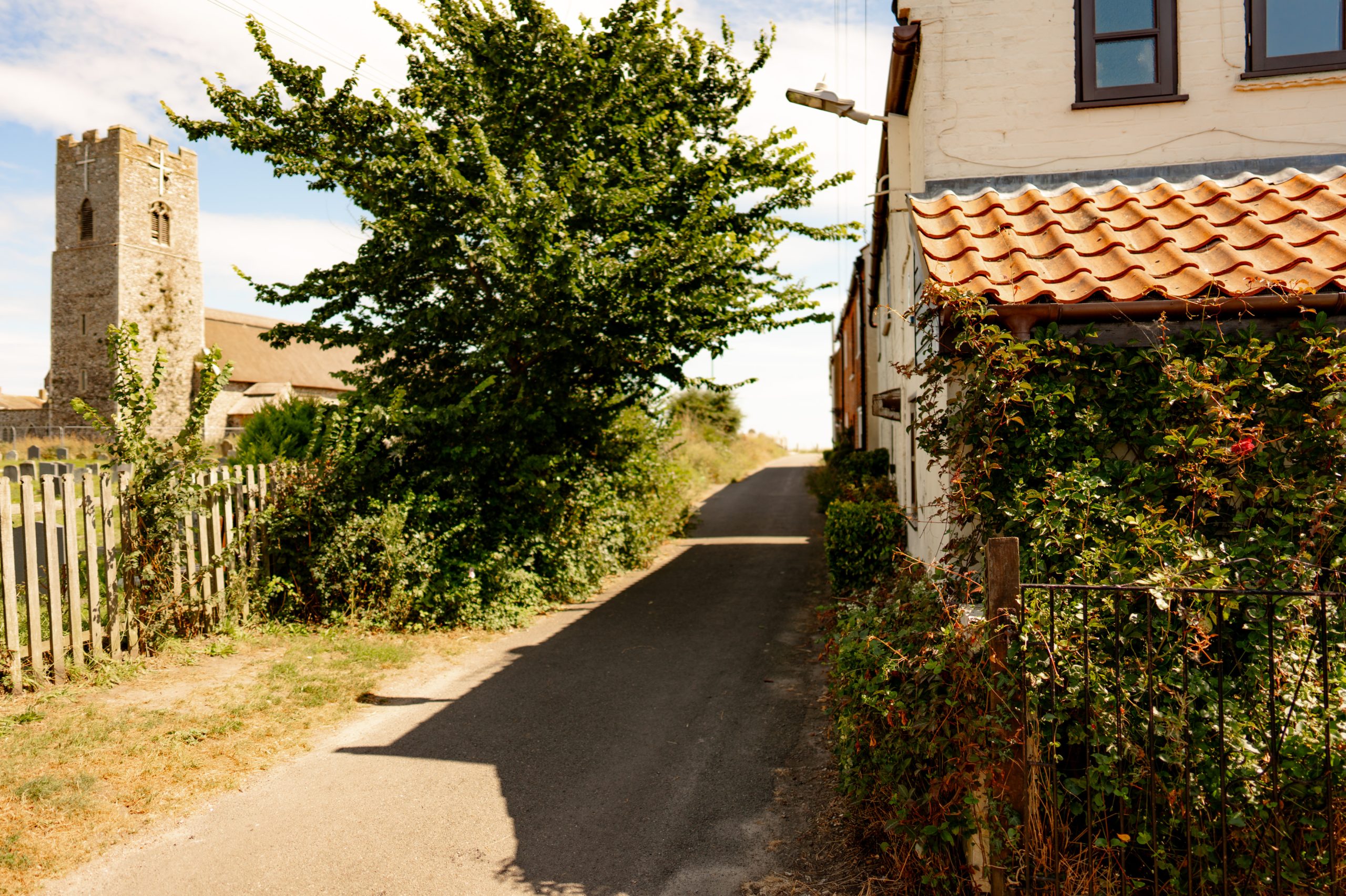 A narrow, sunlit country road flanked by a stone church with a tower on the left and a house with a tile roof on the right. Green foliage lines the road, and a wooden fence runs alongside the church. The sky is clear and bright.