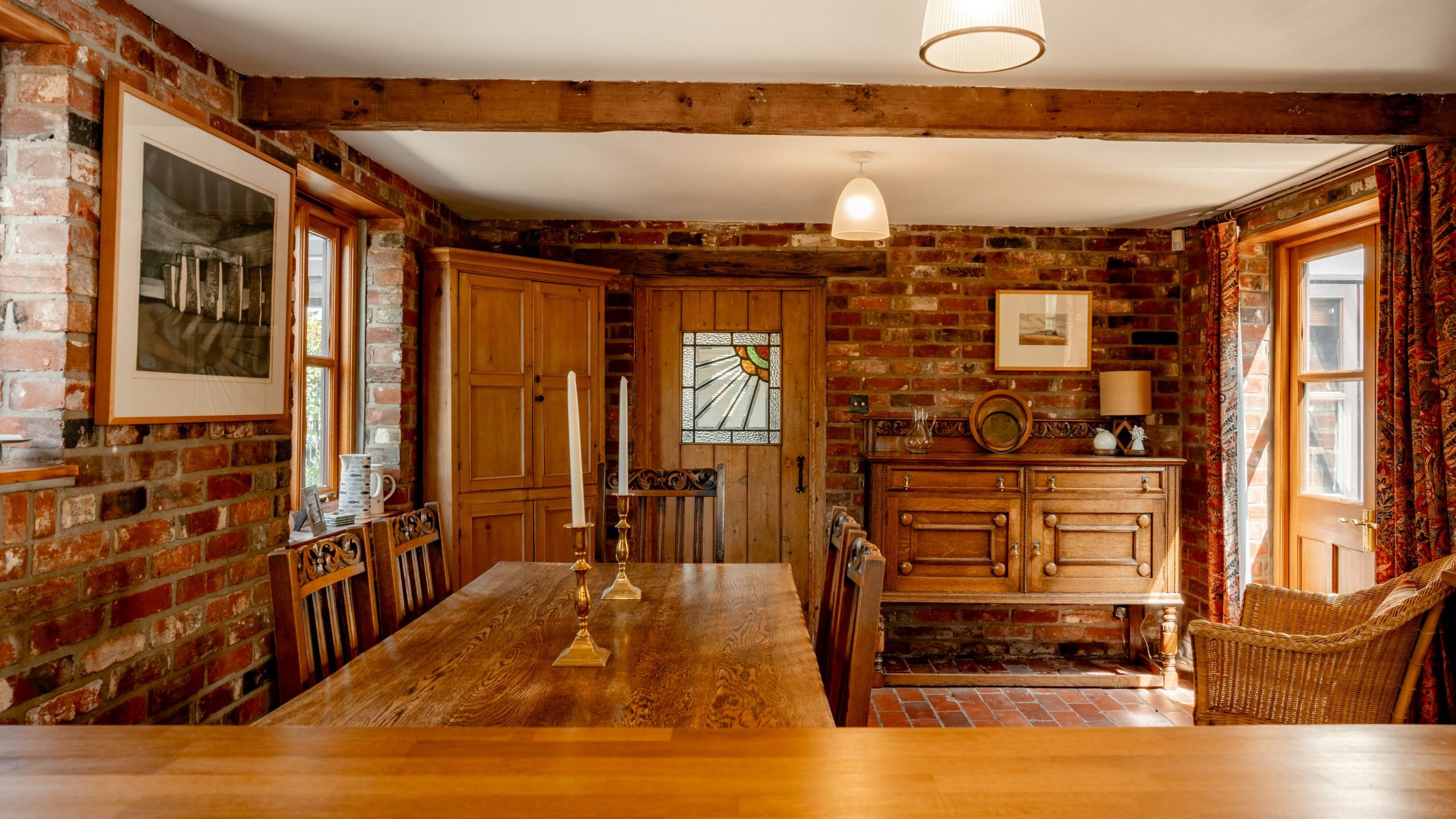 Cozy dining room with brick walls, wooden table and chairs, and a stained glass door. Warm lighting from hanging lamps and natural light through two windows. Wooden furniture and wicker chair add a rustic touch. A framed picture hangs on the left wall.