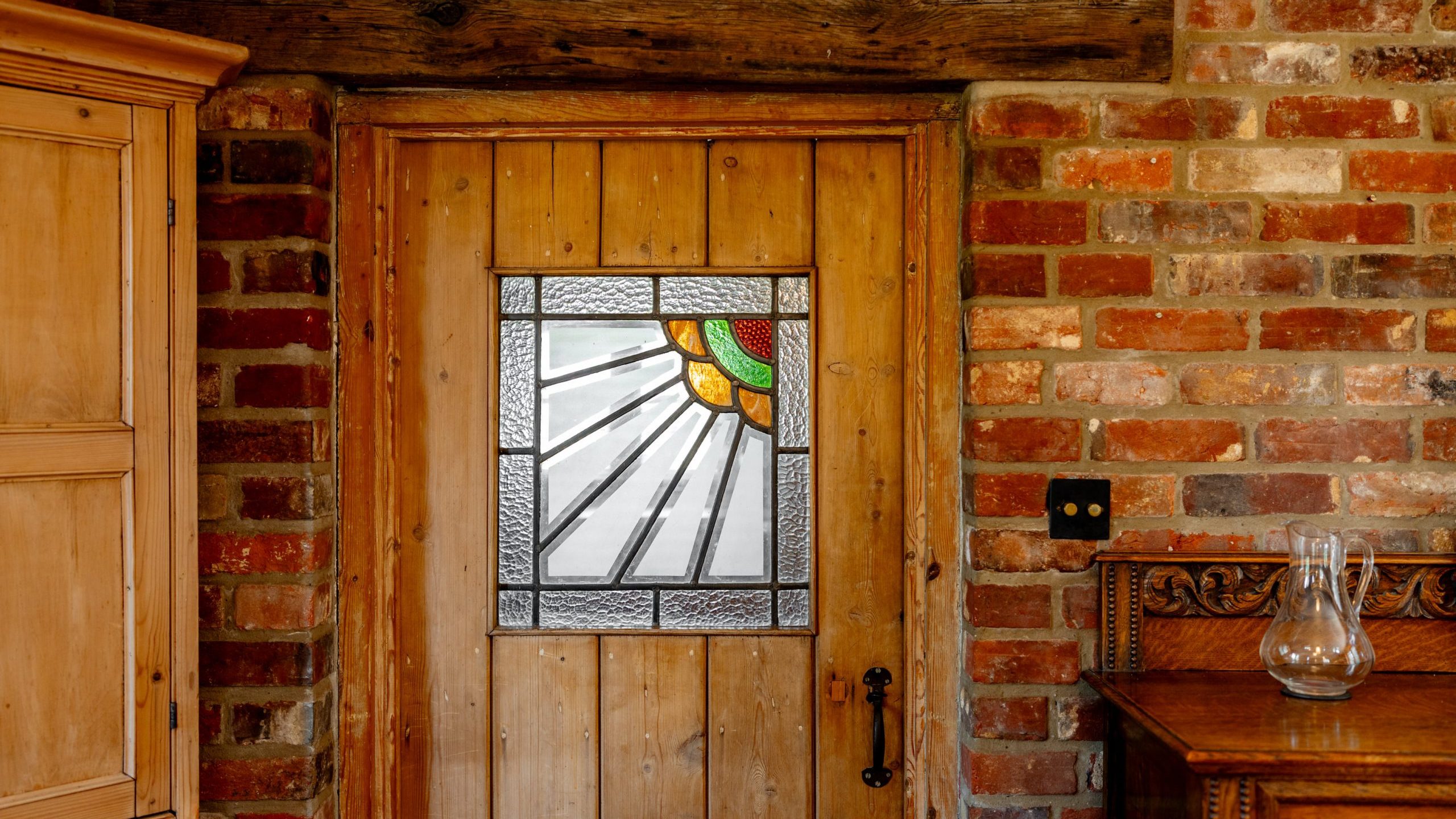 A rustic wooden door with a stained glass window featuring a sunbeam design. The door is set in a brick wall, framed with exposed wooden beams. A wooden cabinet stands to the left, and an ornate wooden dresser with a glass pitcher is on the right.