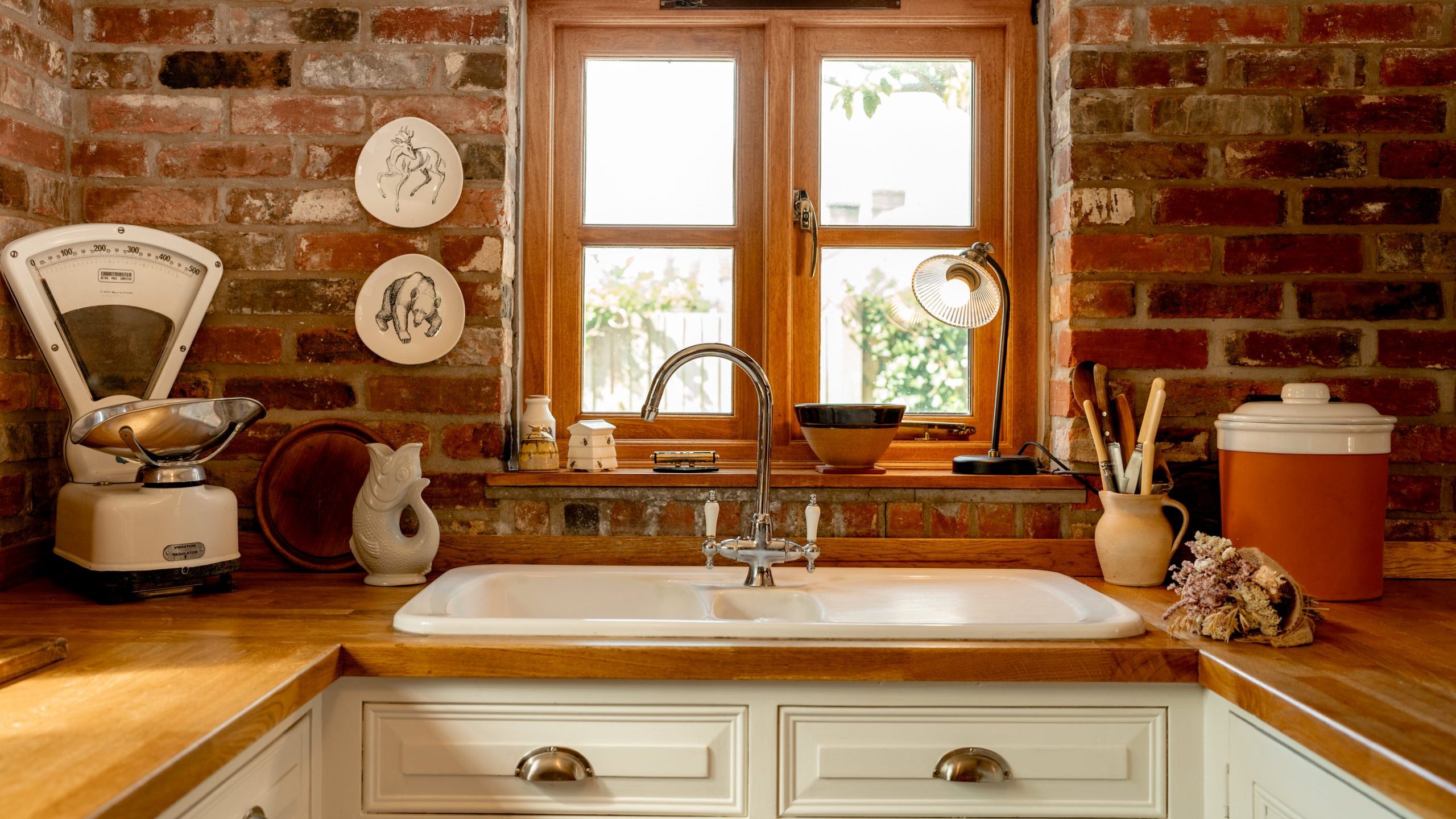 A cozy kitchen with a brick backsplash featuring a window above a white farmhouse sink. On the wooden countertop are a vintage scale, various kitchen utensils in holders, and decorative plates. Natural light streams through the window, illuminating the space.