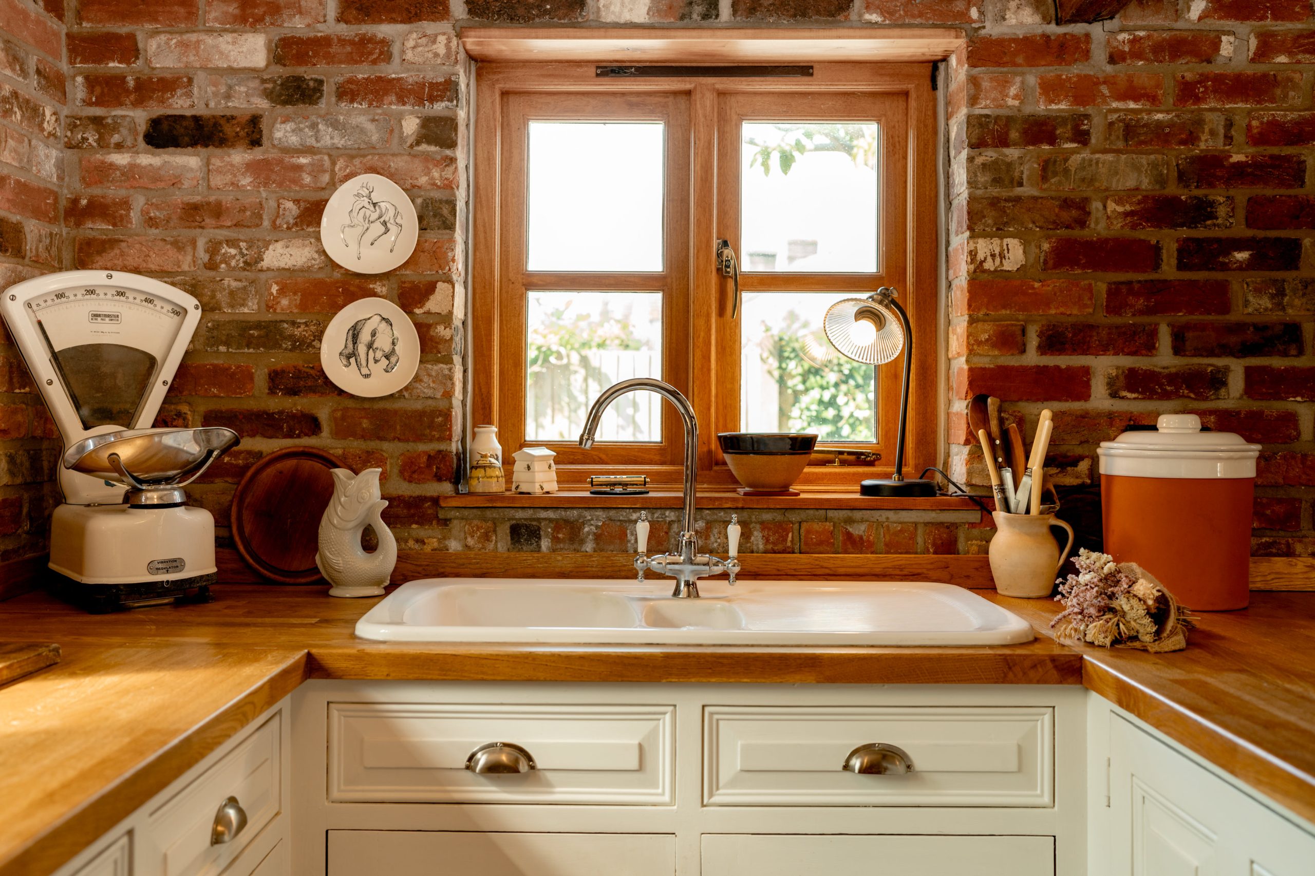 A cozy kitchen with a brick backsplash featuring a window above a white farmhouse sink. On the wooden countertop are a vintage scale, various kitchen utensils in holders, and decorative plates. Natural light streams through the window, illuminating the space.