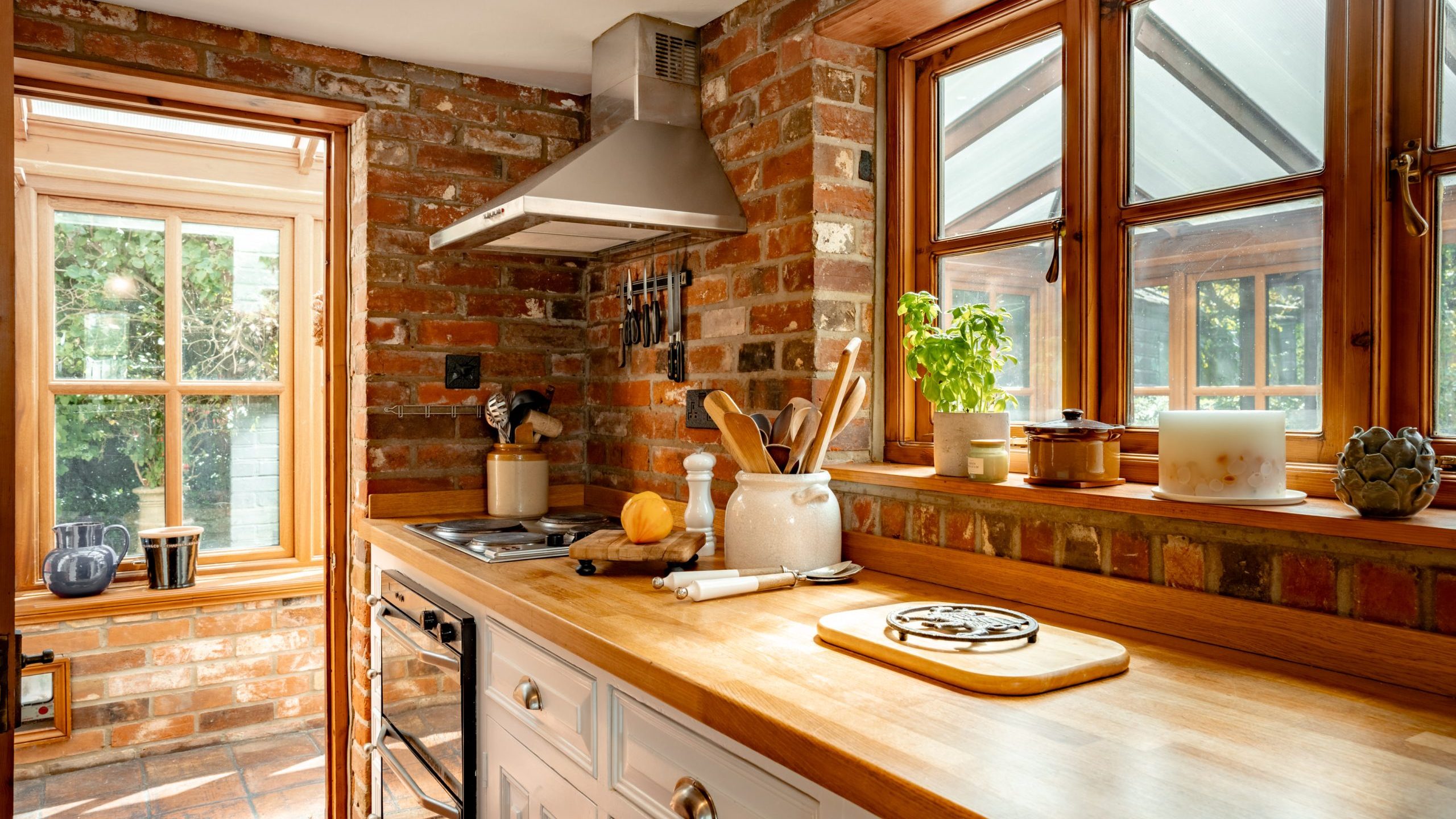 A cozy kitchen with brick walls and wooden countertops. The space is bright, with sunlight coming through two large windows. Kitchen utensils, a plant, and a candle are on the countertop, and a stovetop with a hood is installed near the window.