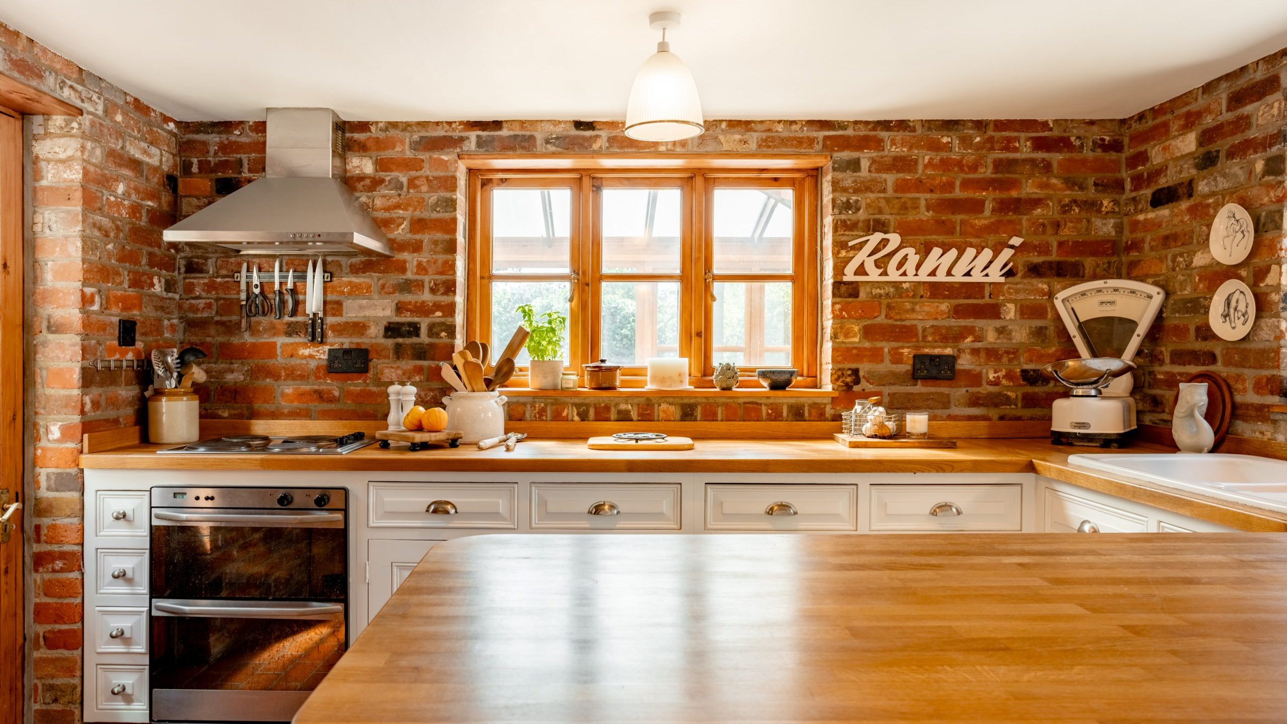 A cozy kitchen features exposed brick walls, wooden countertops, and white cabinetry. Natural light streams in through the window above the sink. Appliances, utensils, and decor items, including a vintage scale and a