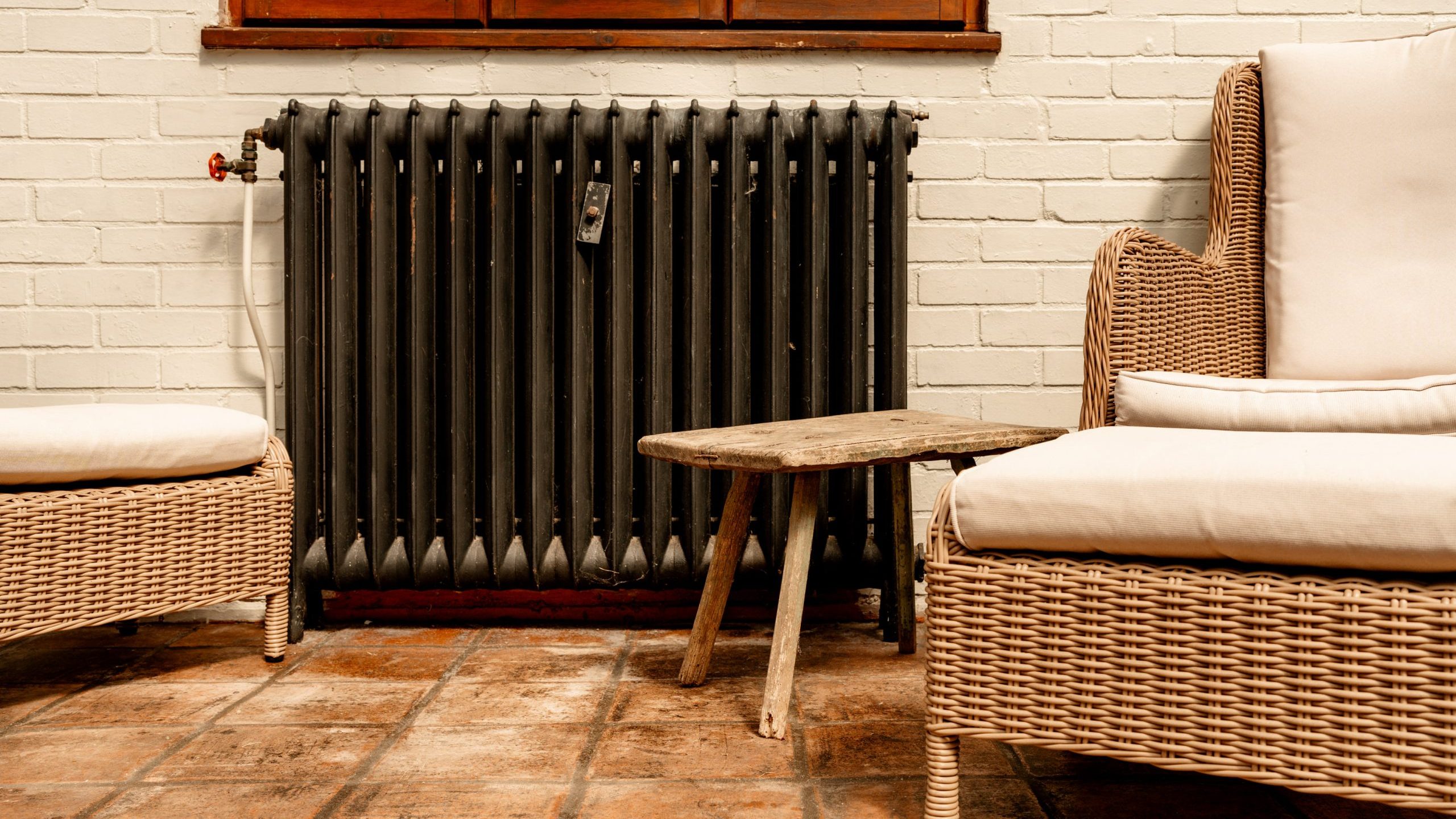 A cozy indoor scene featuring two wicker chairs with cream-colored cushions, a rustic wooden side table in between them, and an old-fashioned black radiator on a white brick wall behind the furniture. The floor is covered with brown, square tiles.