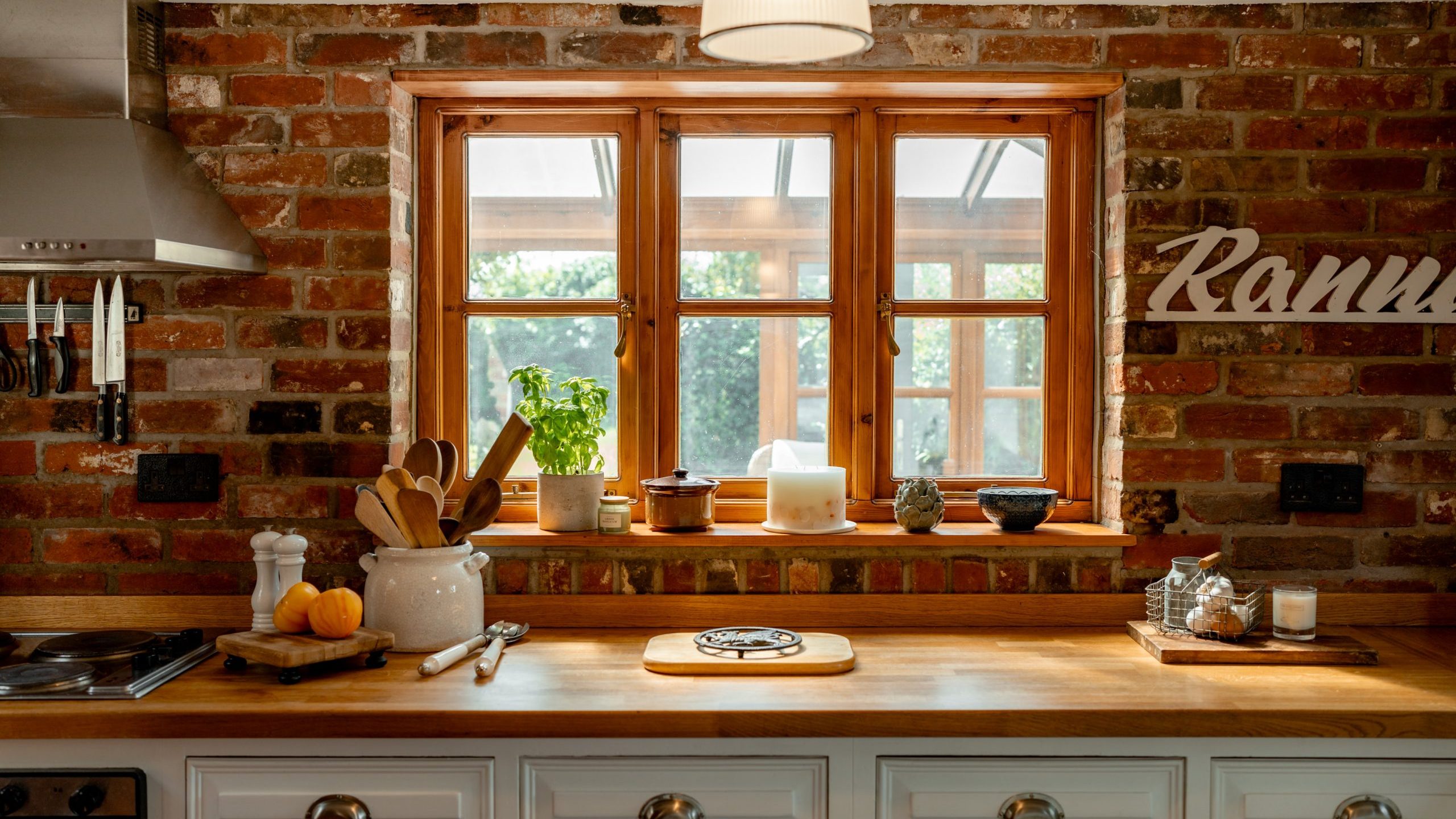 A cozy kitchen with rustic brick walls featuring a wooden countertop. On the countertop are utensils in a ceramic holder, fresh herbs, a cutting board, and a small potted plant by the window. Overhead there's a hanging light fixture. The window has a view of greenery.
