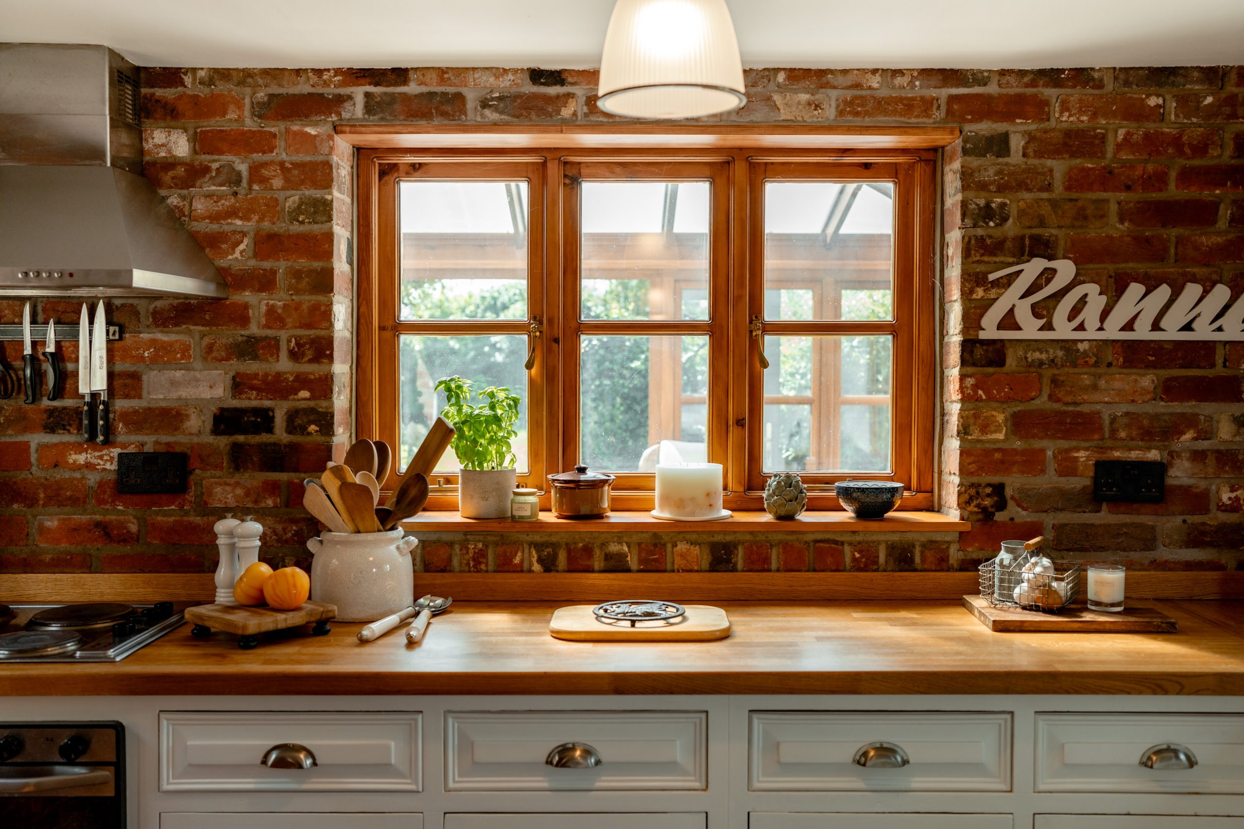 A cozy kitchen with rustic brick walls featuring a wooden countertop. On the countertop are utensils in a ceramic holder, fresh herbs, a cutting board, and a small potted plant by the window. Overhead there's a hanging light fixture. The window has a view of greenery.
