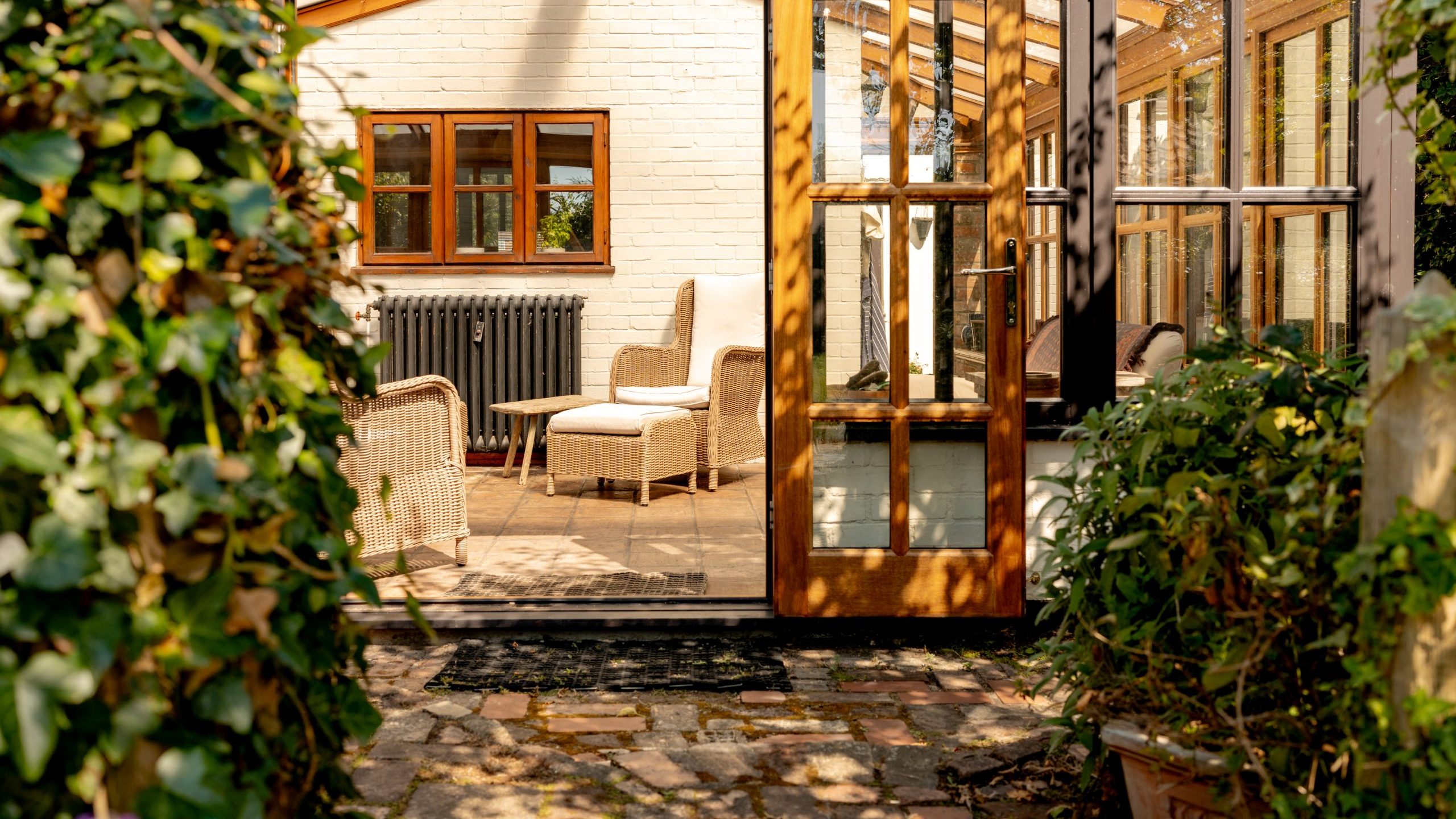 A cozy sunroom with wooden-framed windows and glass doors opening to a lush garden pathway. Inside, wicker chairs with cushions and a small table create a comfortable seating area. Sunlight filters through the foliage, casting dappled shadows inside.