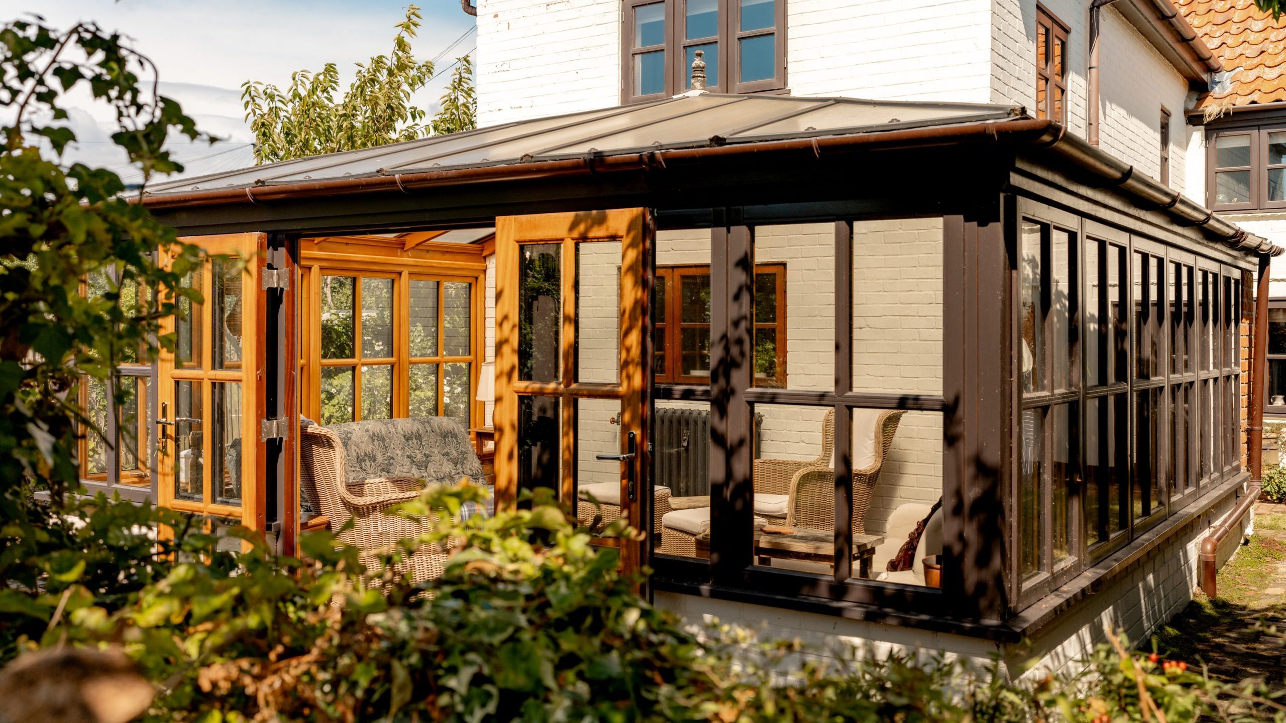 A white house with a large enclosed sunroom featuring many windows and wooden frames. Inside, there are wicker furniture pieces and potted plants. The sunroom is surrounded by greenery, with bushes and trees partially visible in the foreground.
