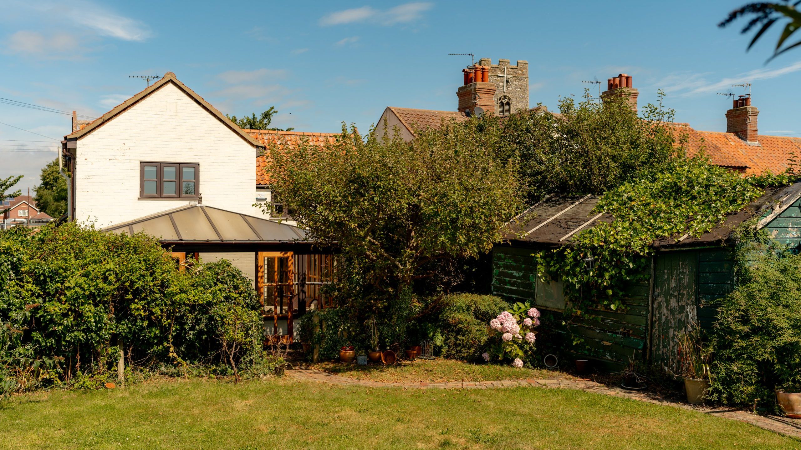 A sunny backyard featuring a white house with a brown roof and large windows, surrounded by lush greenery and trees. Adjacent to the house is a small gazebo. A green wooden shed and plants with pink flowers are also visible in the yard.