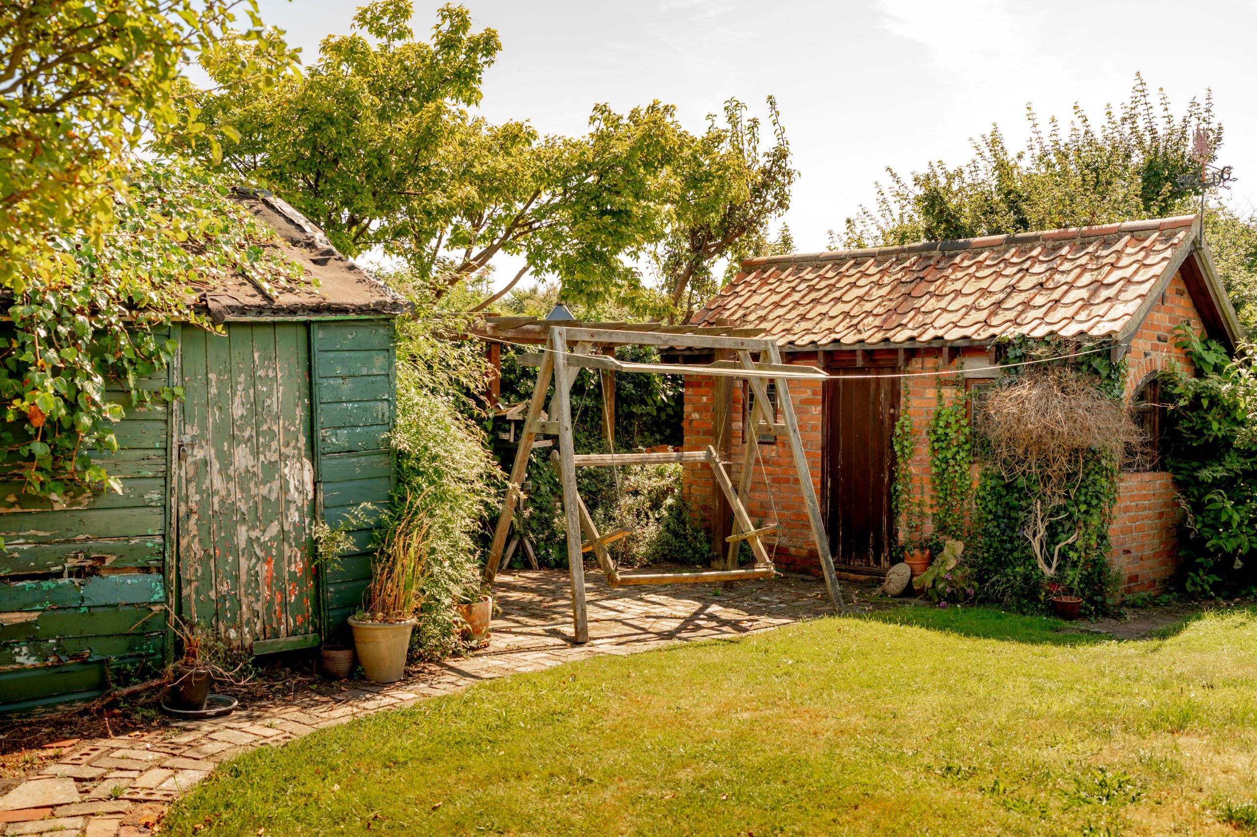 A rustic backyard scene with a weathered green shed on the left, a wooden swing set in the center, and a small brick building with a tiled roof on the right. Lush greenery surrounds the area, including potted plants and trees, with a neatly mowed lawn in the foreground.