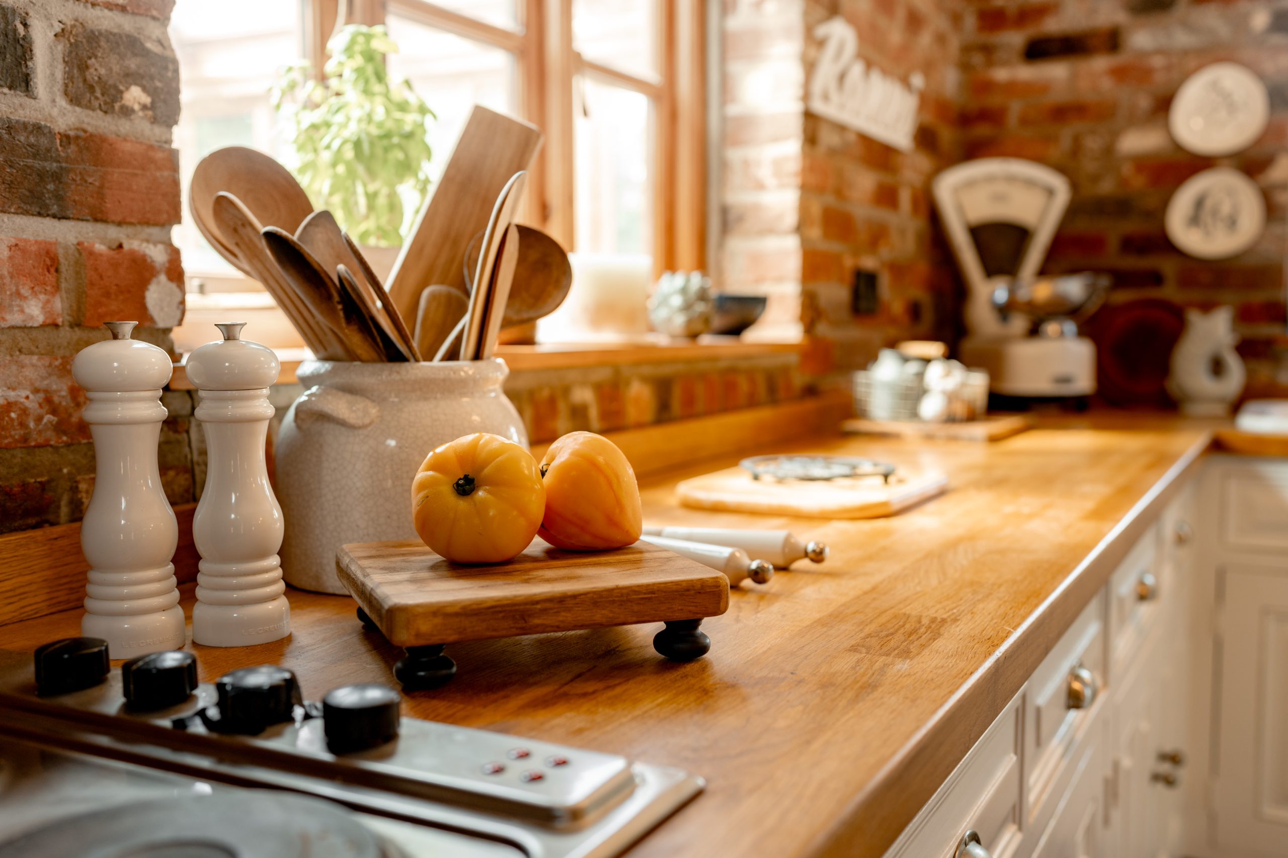 A rustic kitchen with a wooden countertop featuring two yellow tomatoes on a small cutting board. A white ceramic container holding wooden utensils, salt and pepper shakers, and a vintage scale are seen in the background. Sunlight streams in through the window.