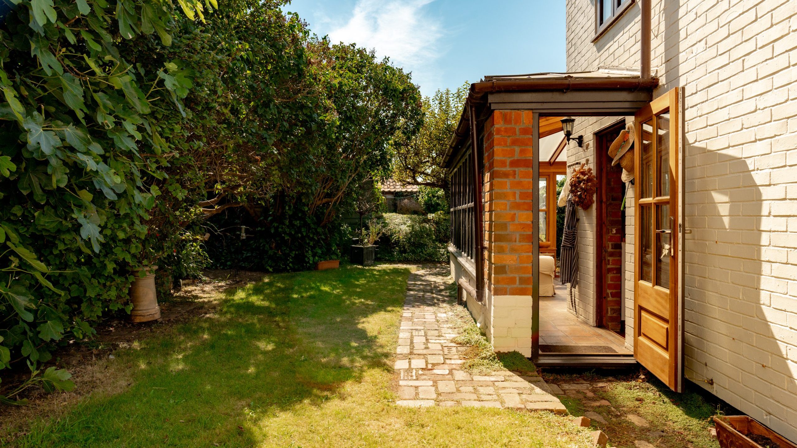 A sunny garden with a grassy lawn and a paved stone pathway leading to a partially brick, partially white-painted house. The house has an open wooden door, with a narrow outdoor corridor and large green leafy trees providing shade on the left.