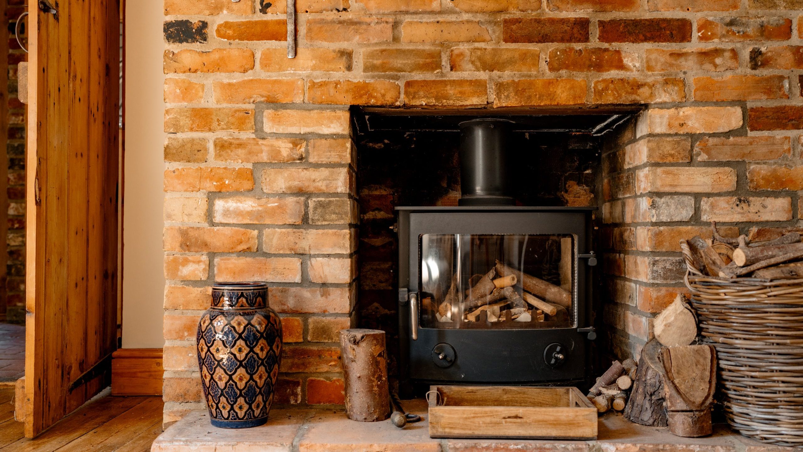 A cozy room with a brick fireplace features a black wood-burning stove filled with logs. A large decorative vase and a rustic log holder sit nearby, and a wooden door is open, leading to another room. The warm tones of the bricks and wooden floor add to the inviting atmosphere.