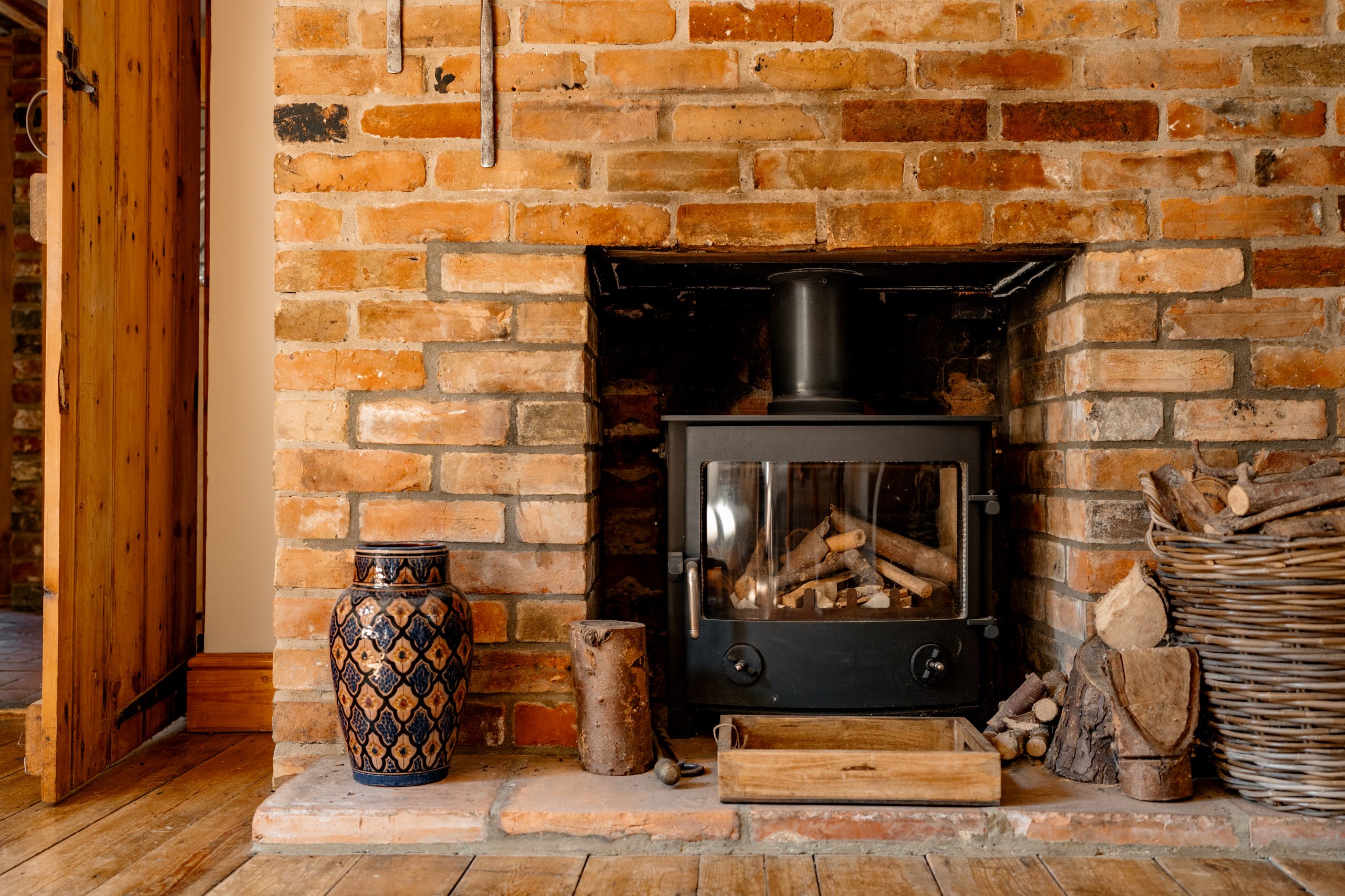 A cozy room with a brick fireplace features a black wood-burning stove filled with logs. A large decorative vase and a rustic log holder sit nearby, and a wooden door is open, leading to another room. The warm tones of the bricks and wooden floor add to the inviting atmosphere.