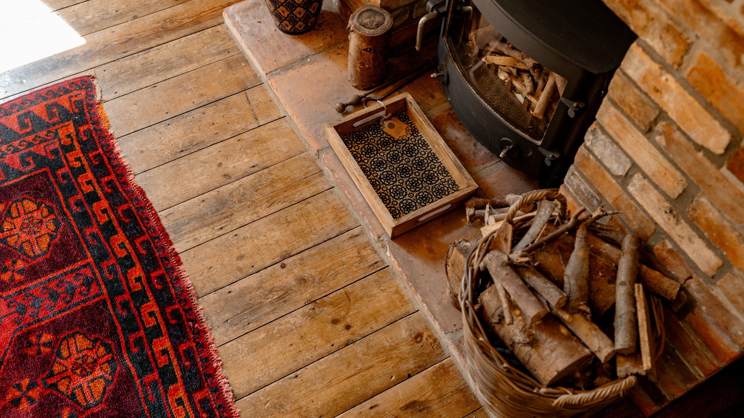 Sunlit wooden floor by a brick fireplace with a wood stove, basket of firewood, small decorative objects, and an ornate red rug. A cozy ambiance with warm hues and rustic elements.