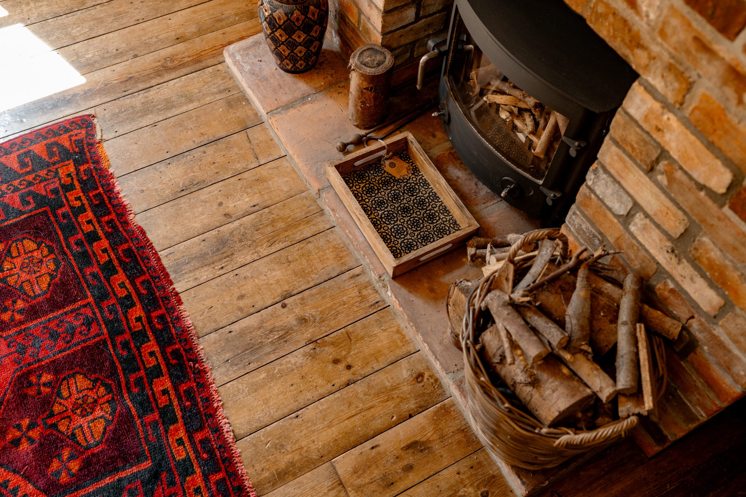 Sunlit wooden floor by a brick fireplace with a wood stove, basket of firewood, small decorative objects, and an ornate red rug. A cozy ambiance with warm hues and rustic elements.