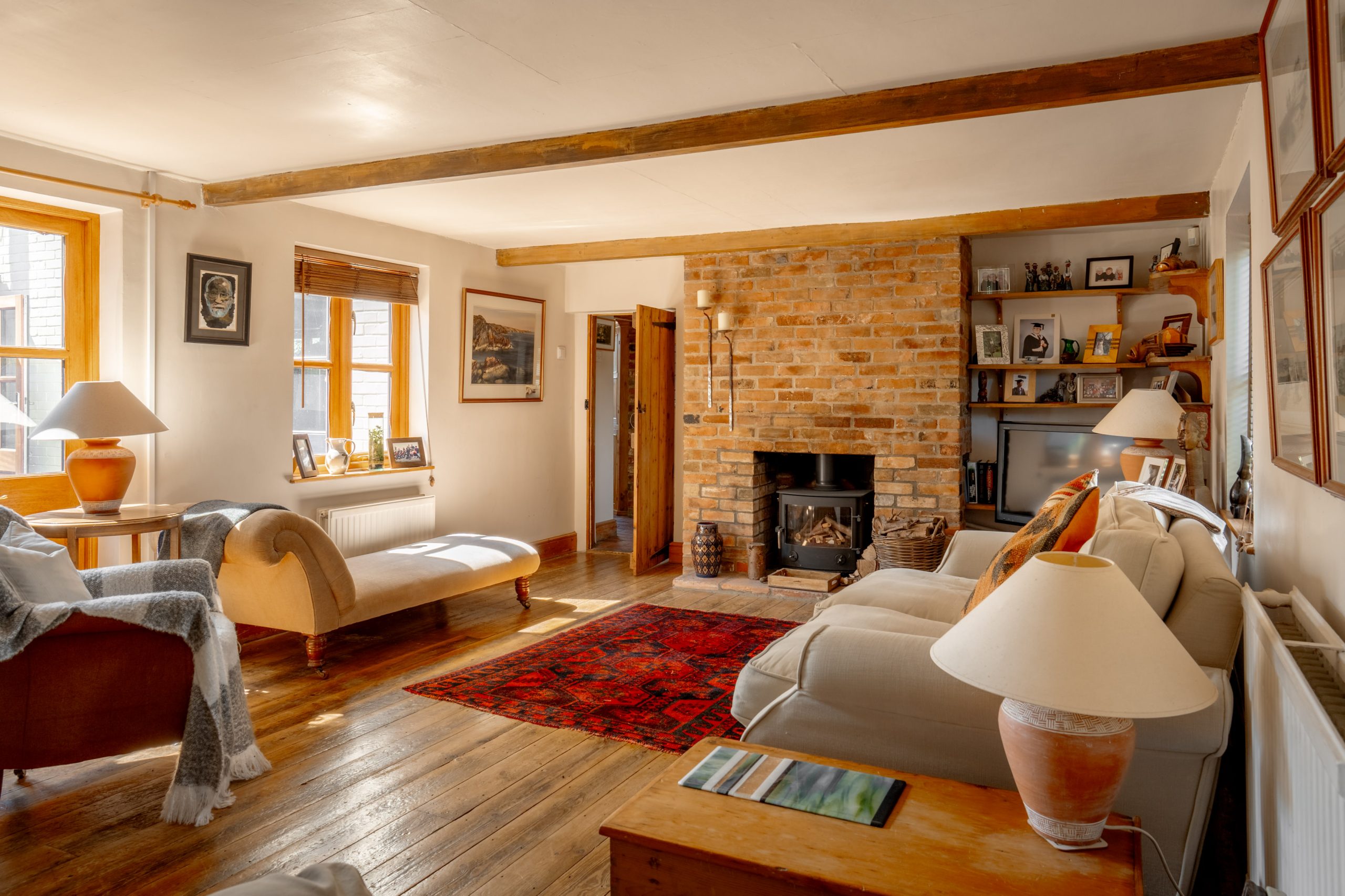 A cozy living room with wooden floors, exposed wooden ceiling beams, and a brick fireplace in the center. There are comfortable sofas, a cushioned bench, and various framed pictures on the walls and shelves. Sunlight streams through the windows.