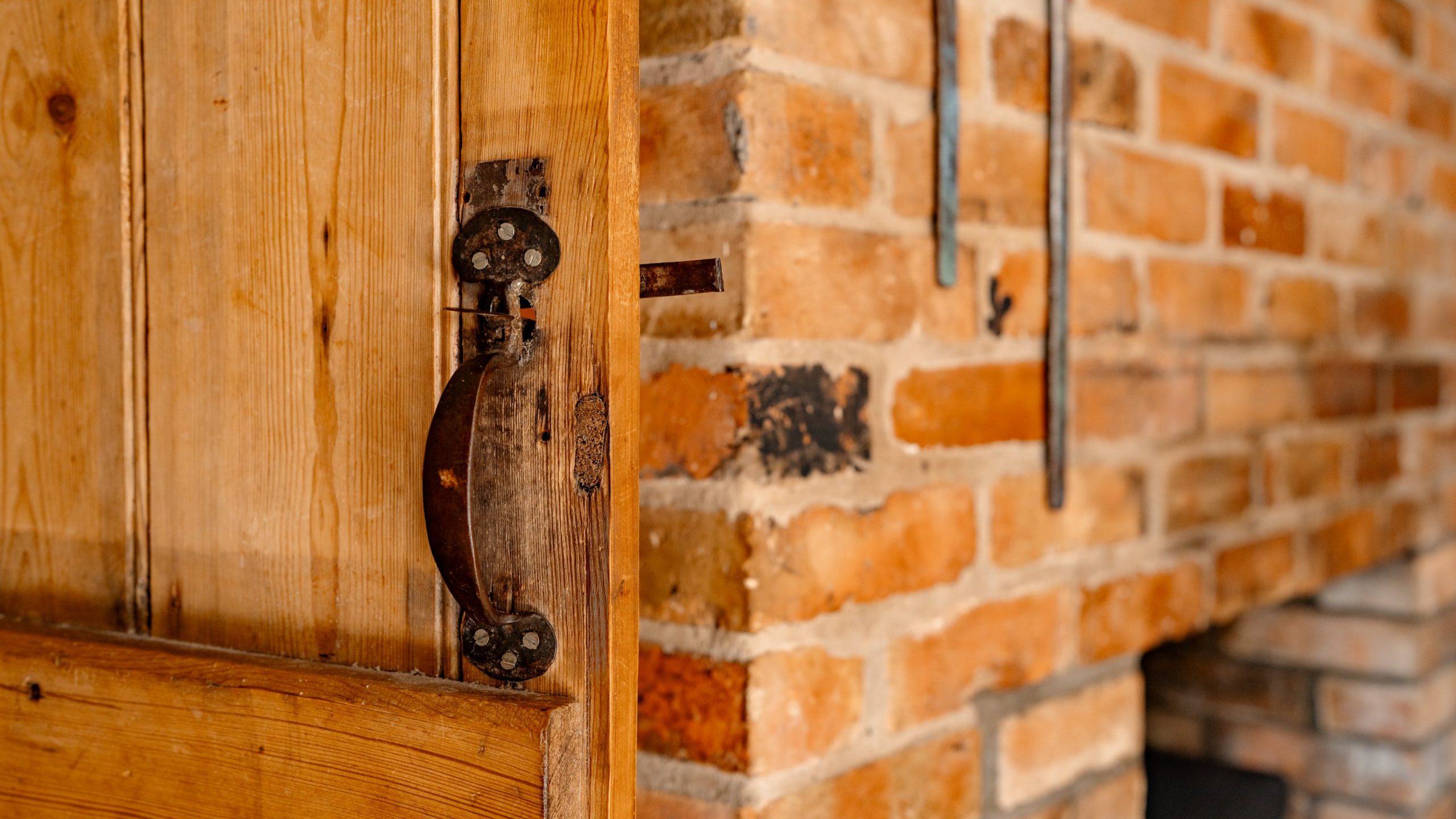 A close-up of a rustic wooden door with a metal latch, slightly ajar, revealing a brick wall and part of a fireplace in the background. The scene exudes a warm, cozy atmosphere.