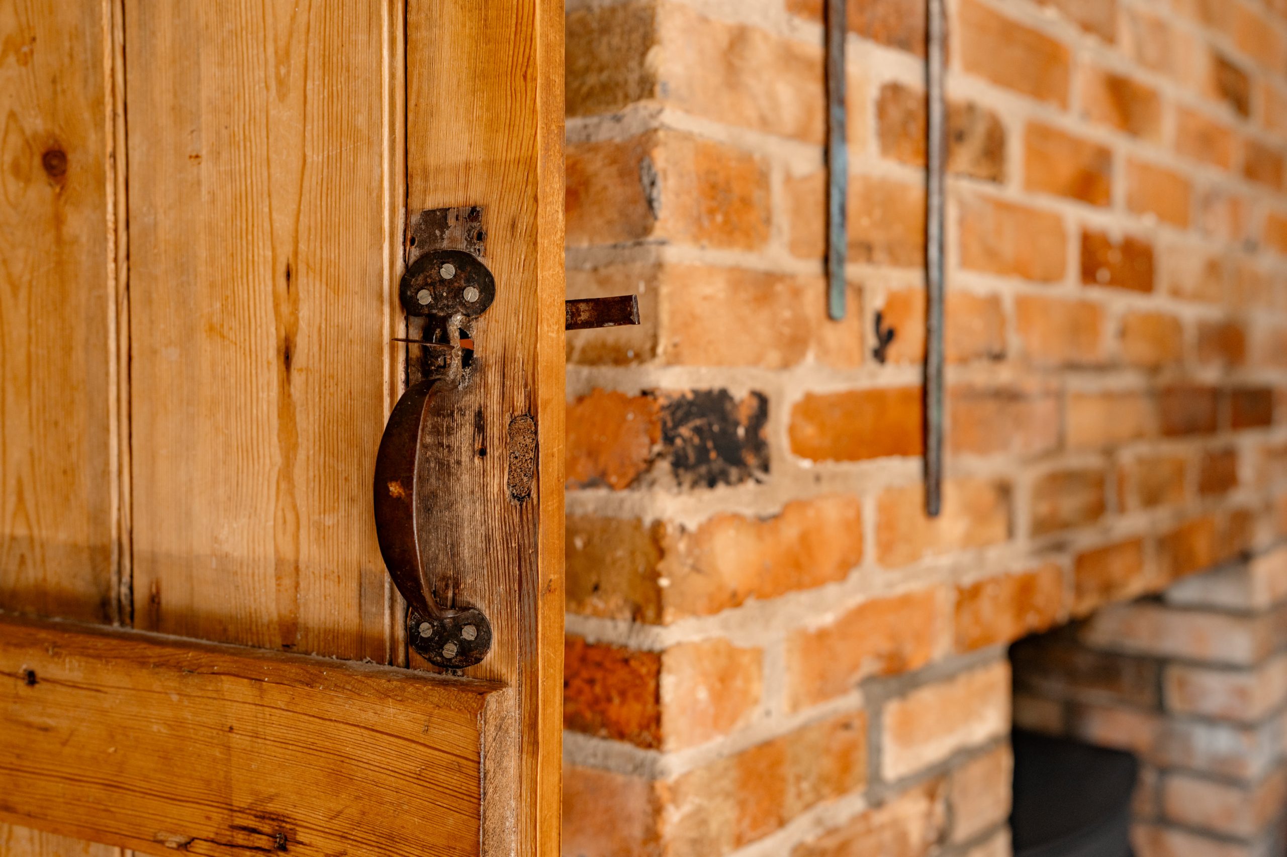 A close-up of a rustic wooden door with a metal latch, slightly ajar, revealing a brick wall and part of a fireplace in the background. The scene exudes a warm, cozy atmosphere.