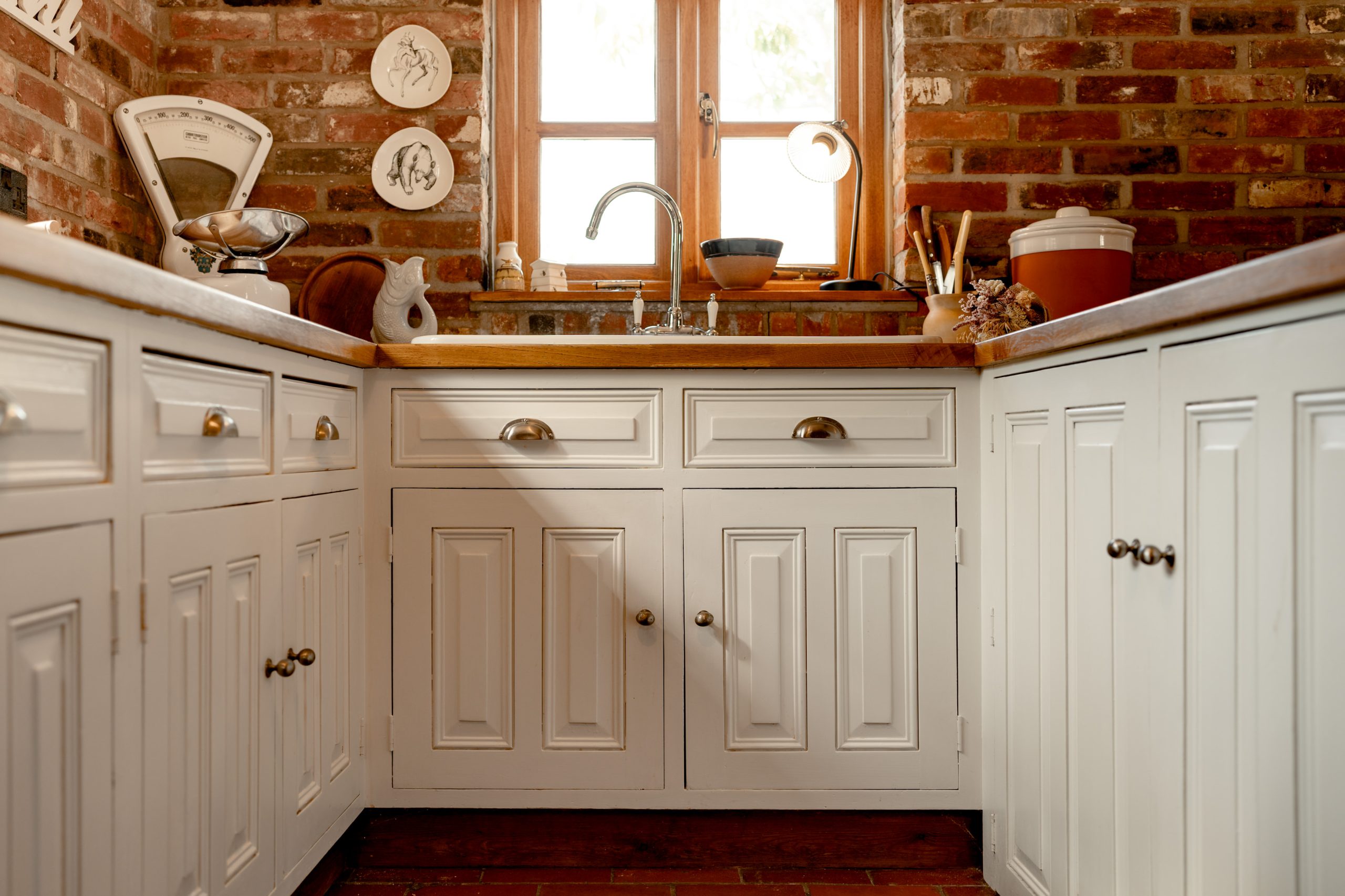 A cozy kitchen with white cabinets, wooden countertops, and a central sink under a small, two-paned window. The brick walls add rustic charm. Kitchen items and decor, including a bowl and kitchen scale, are arranged neatly on the countertops and shelves.