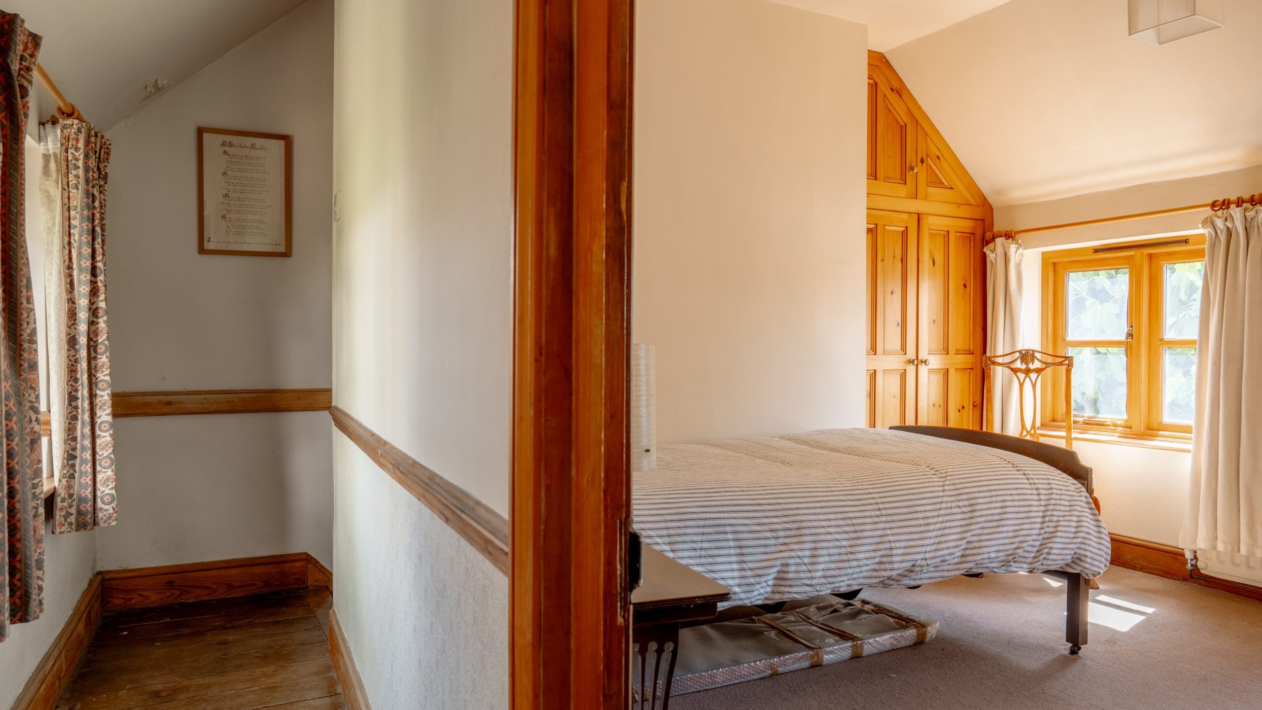A bright, rustic bedroom with wooden trim and a large window. The bed has a striped comforter, with a wooden headboard and footboard. On the left, a wooden hallway features a curtained window. The room is lit by natural light streaming through the windows.