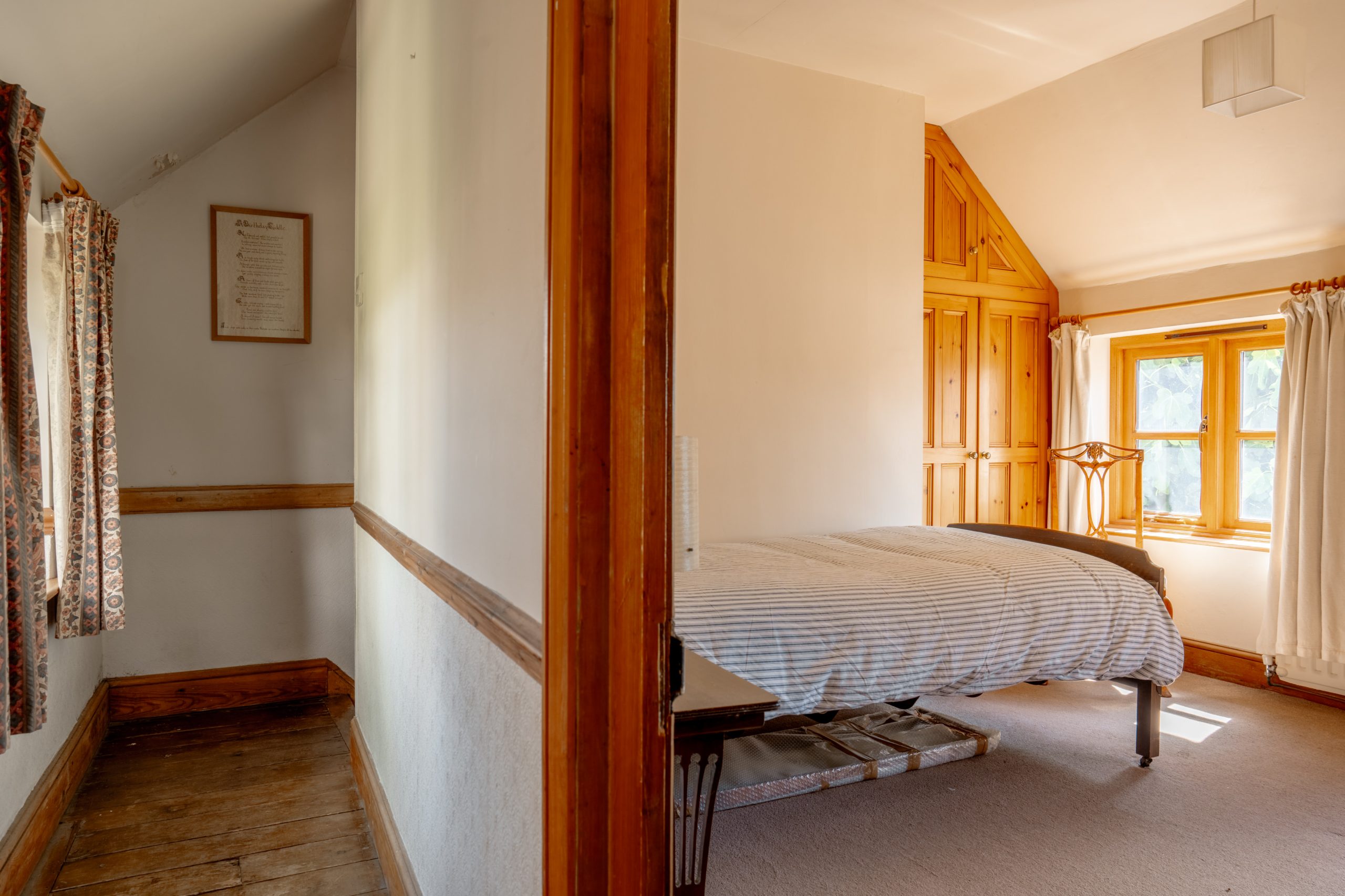 A bright, rustic bedroom with wooden trim and a large window. The bed has a striped comforter, with a wooden headboard and footboard. On the left, a wooden hallway features a curtained window. The room is lit by natural light streaming through the windows.