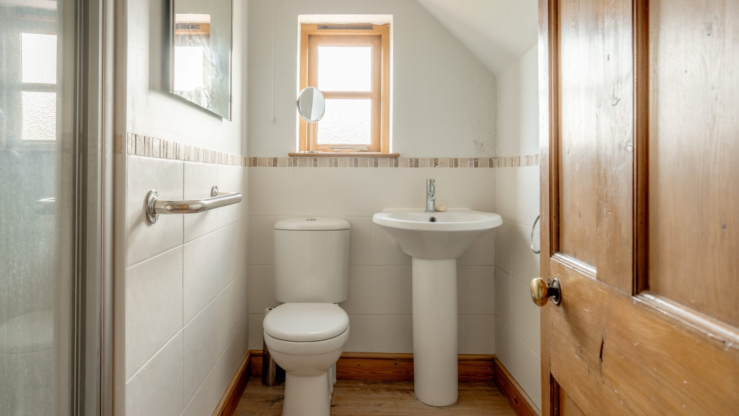 A small bathroom with a wooden door, white toilet, and matching pedestal sink. The walls are tiled halfway up with white tiles and a decorative border. A small window above the toilet allows natural light in. There's also a small mirror mounted on the wall.