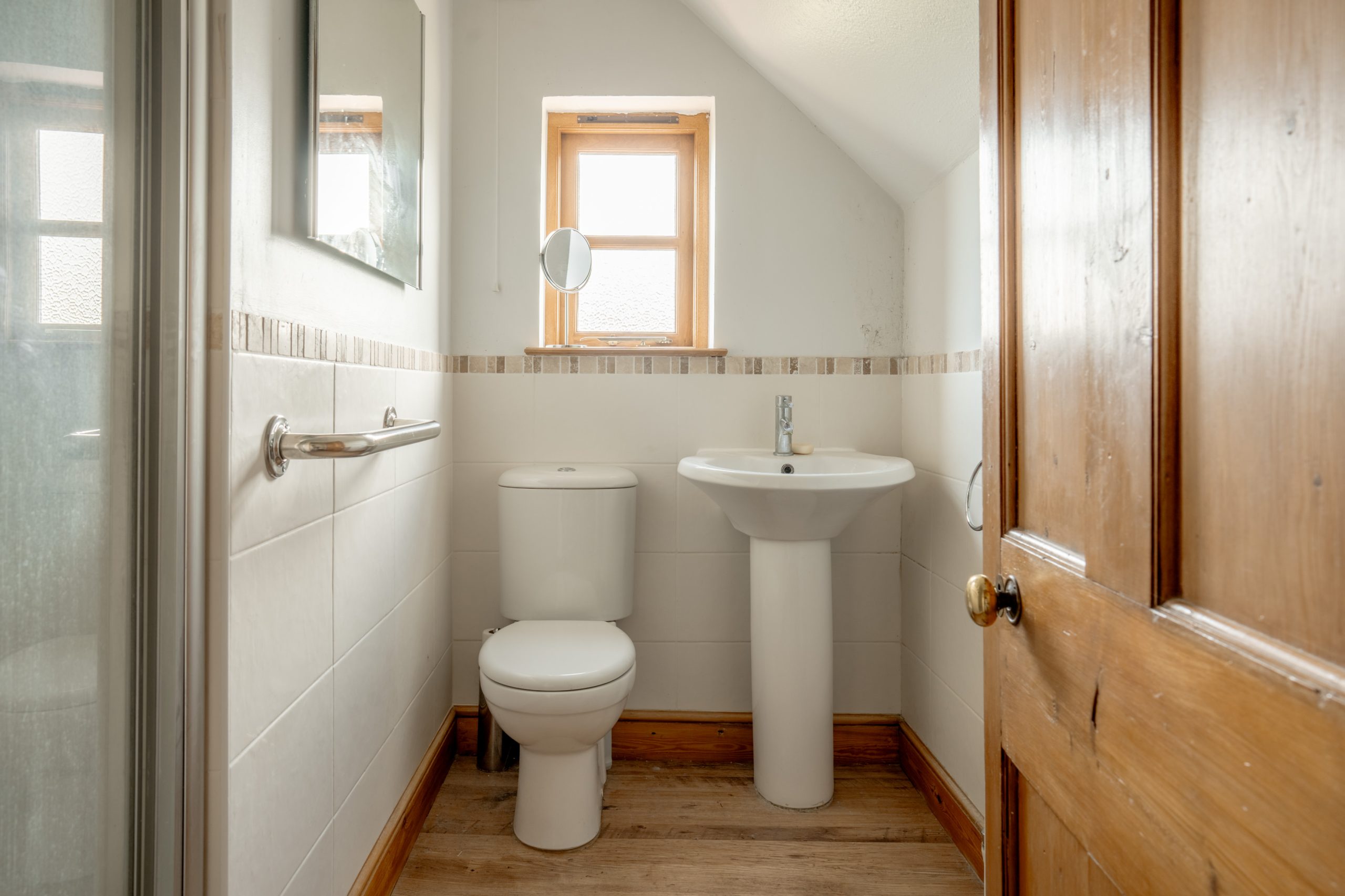 A small bathroom with a wooden door, white toilet, and matching pedestal sink. The walls are tiled halfway up with white tiles and a decorative border. A small window above the toilet allows natural light in. There's also a small mirror mounted on the wall.