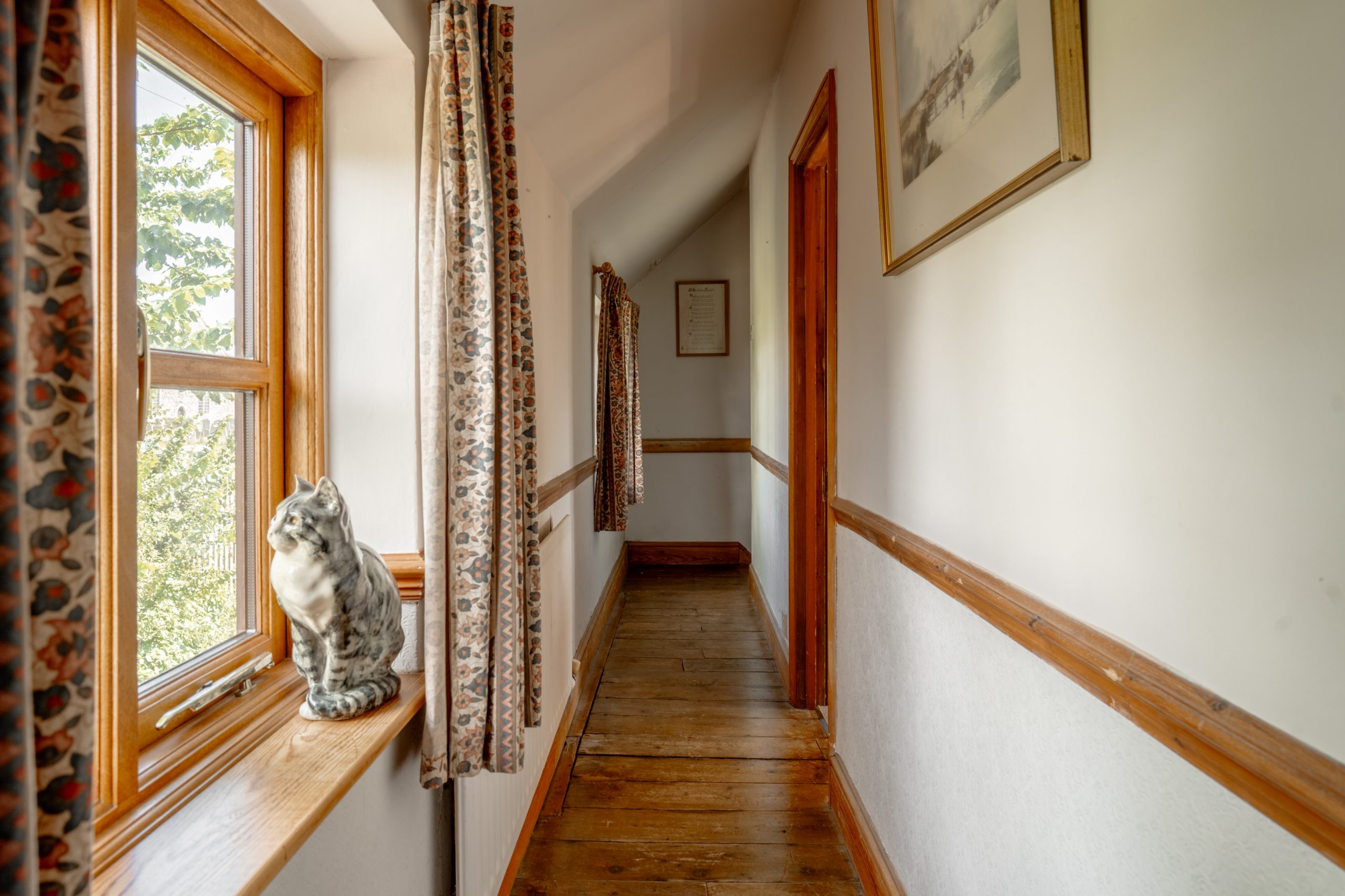 A narrow hallway with a wooden floor, white walls, and framed pictures. A window with patterned curtains on the left allows sunlight to enter. A decorative ceramic cat sits on the window sill. The hallway leads to a wooden door at the far end.