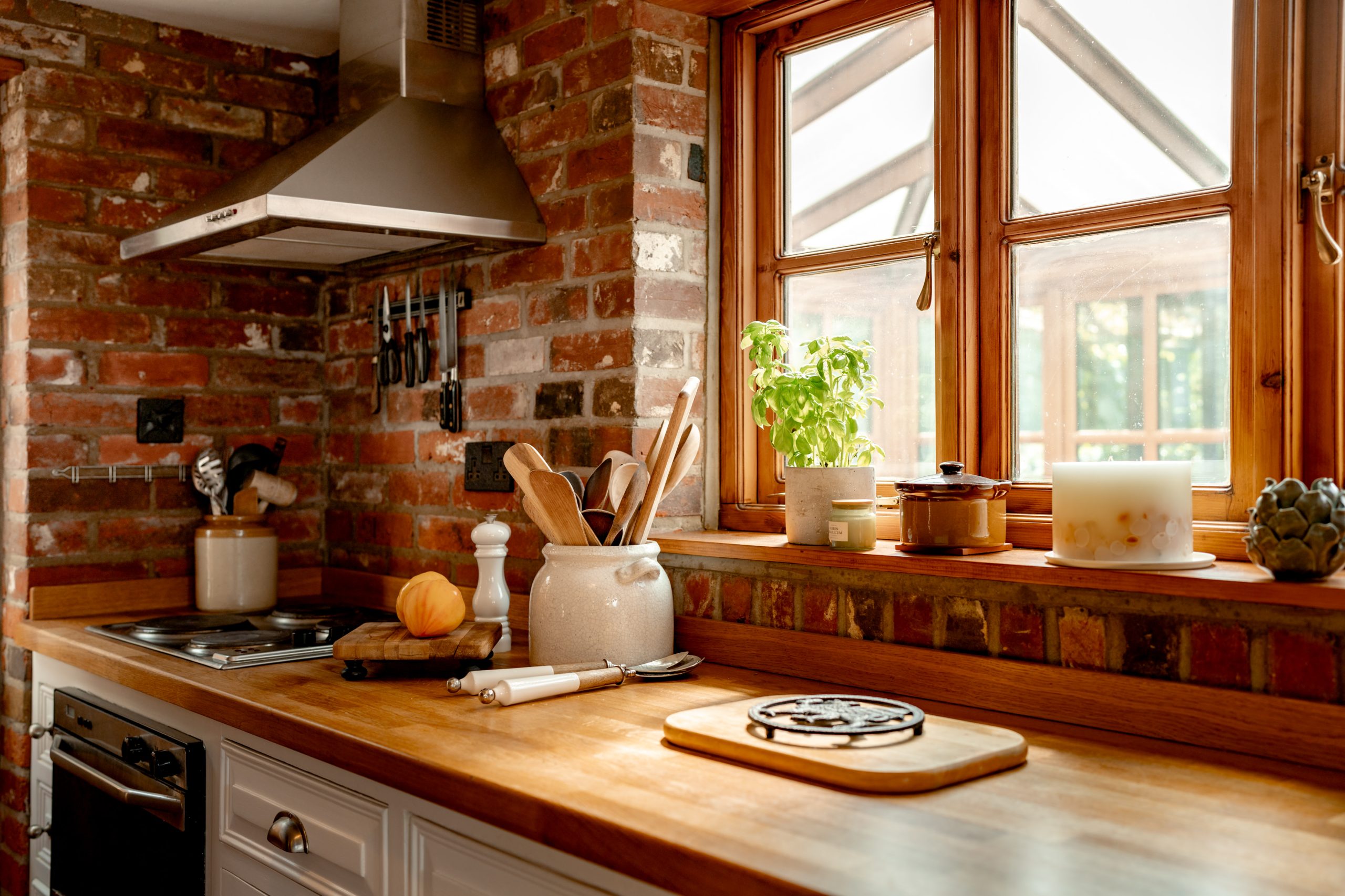 A cozy kitchen featuring rustic brick walls and wooden countertops. The counter holds various utensils, a potted plant, a candle, and a cutting board. A window with wooden frames lets in natural light, and a stainless steel range hood is above the stove.