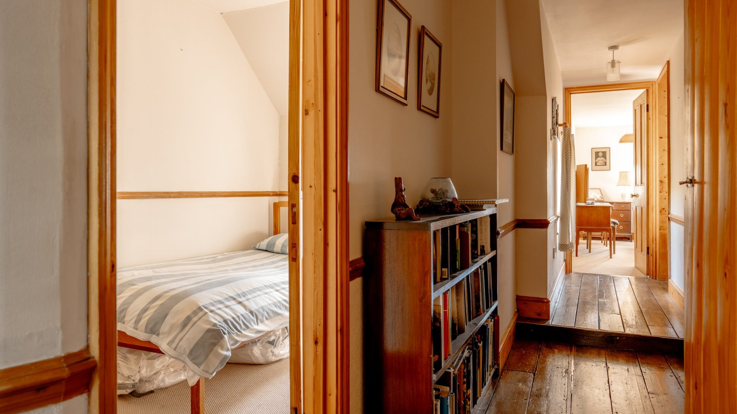A hallway with wooden floors and a bookshelf leads to an open door on the right, revealing a cozy bedroom with a striped bedspread. Another room with a desk and lamp is visible further down the hallway through an open door. The walls are adorned with framed pictures.