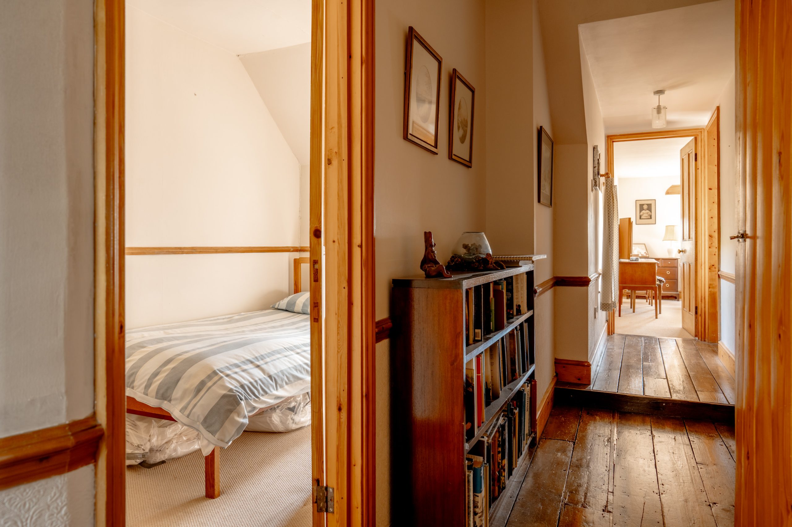 A hallway with wooden floors and a bookshelf leads to an open door on the right, revealing a cozy bedroom with a striped bedspread. Another room with a desk and lamp is visible further down the hallway through an open door. The walls are adorned with framed pictures.