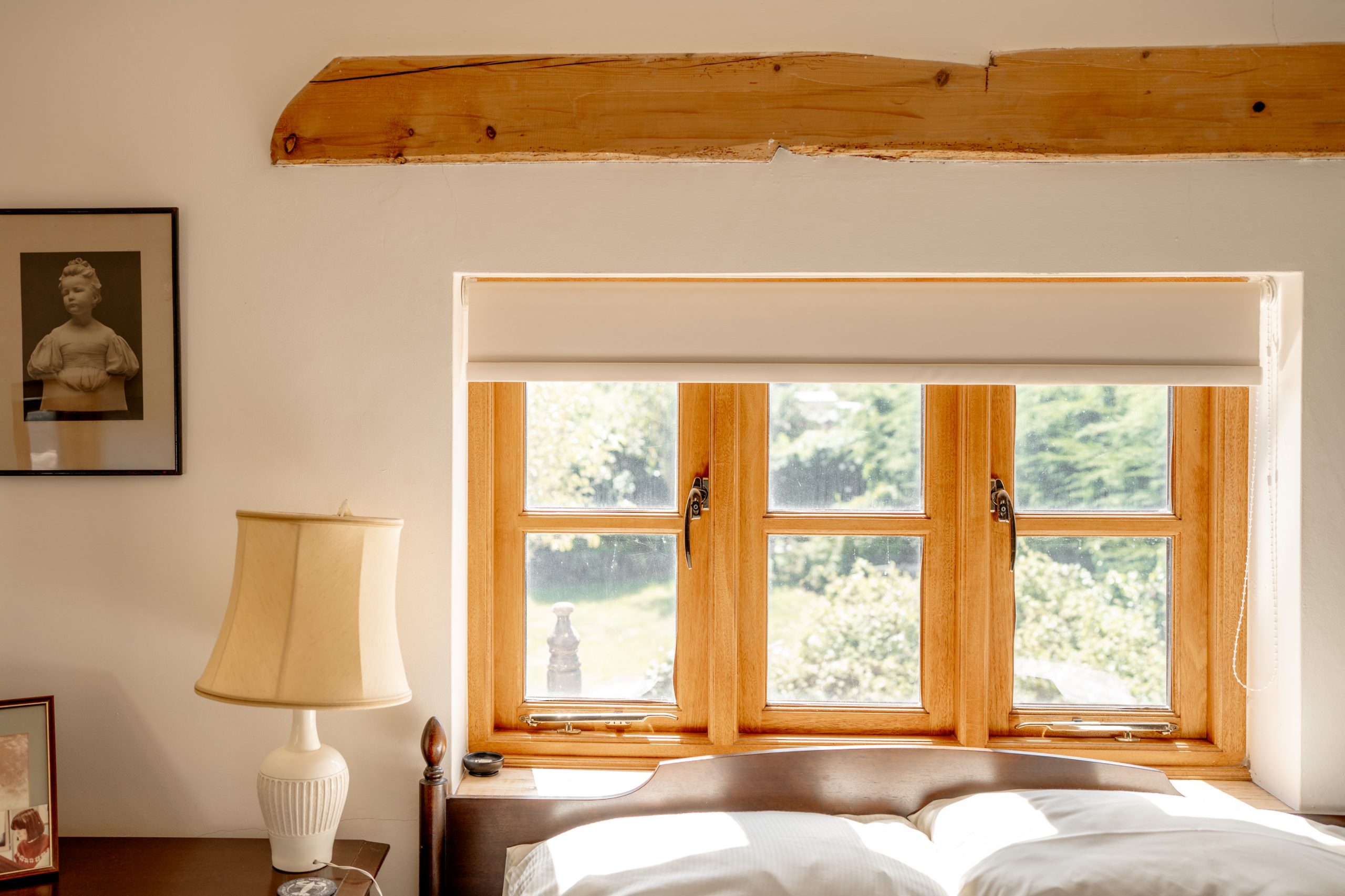 A cozy bedroom features a wooden beam above a window with sunlight streaming in. The window has a white roller shade partially open, revealing a view of greenery outside. A bedside table holds a beige lampshade and framed pictures, including a black-and-white portrait.