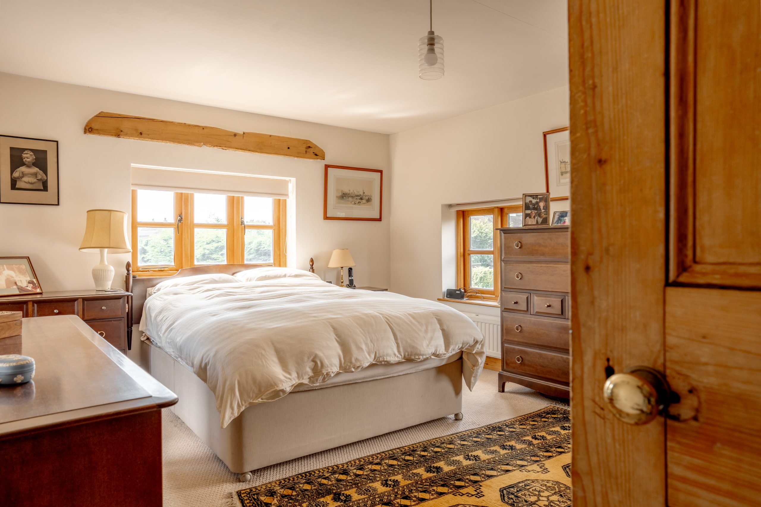 A cozy bedroom with a double bed in the center, covered with a white duvet. The room features wooden furniture, including dressers and side tables, and is softly lit by a combination of natural light from the windows and a ceiling pendant light. Various paintings adorn the walls.