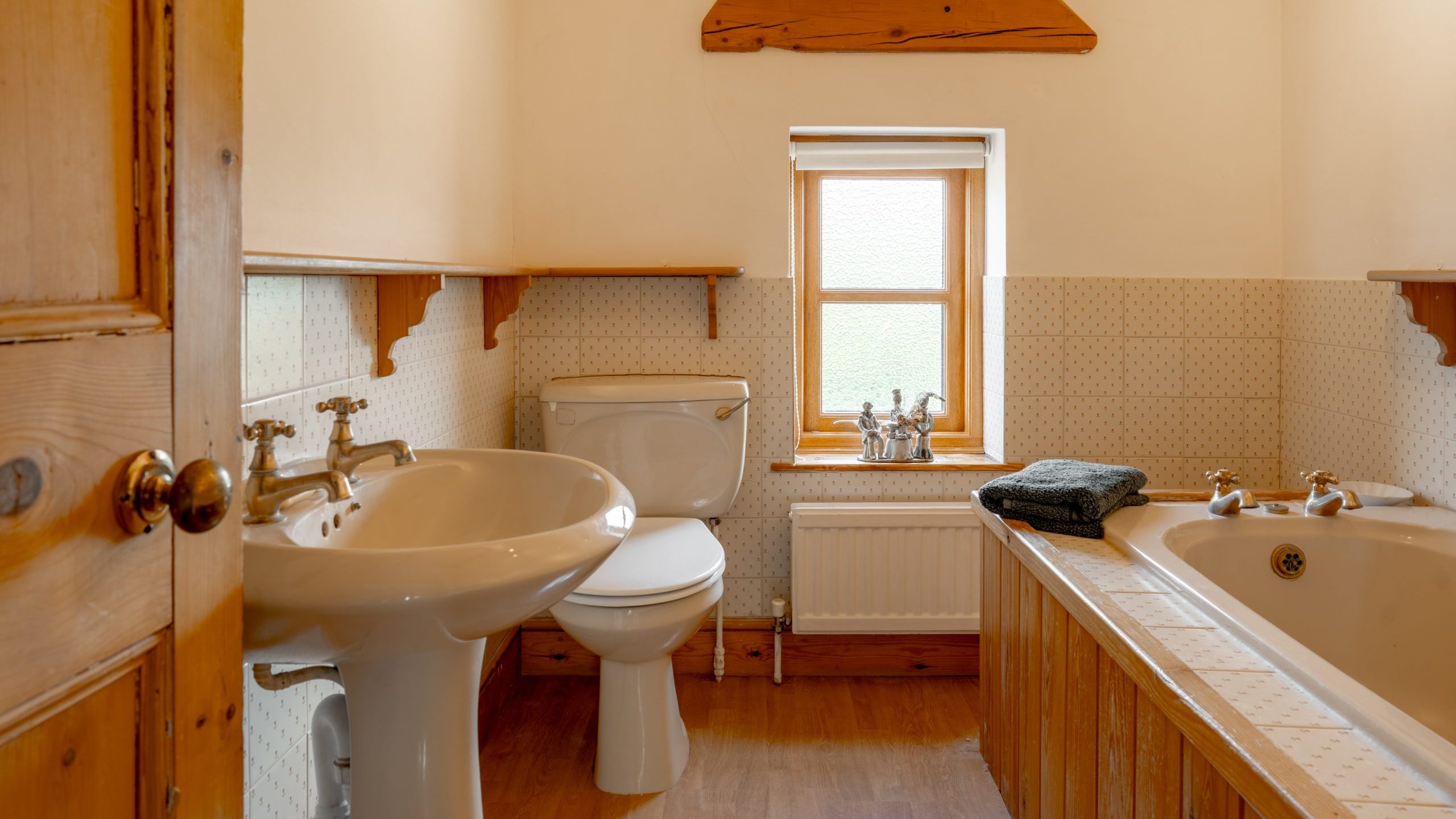 A cozy bathroom features a white sink, a white toilet, and a wooden-framed bathtub. The room is decorated with wooden accents and has a small frosted window. Two folded towels are placed on the edge of the bathtub. The floor is wooden.
