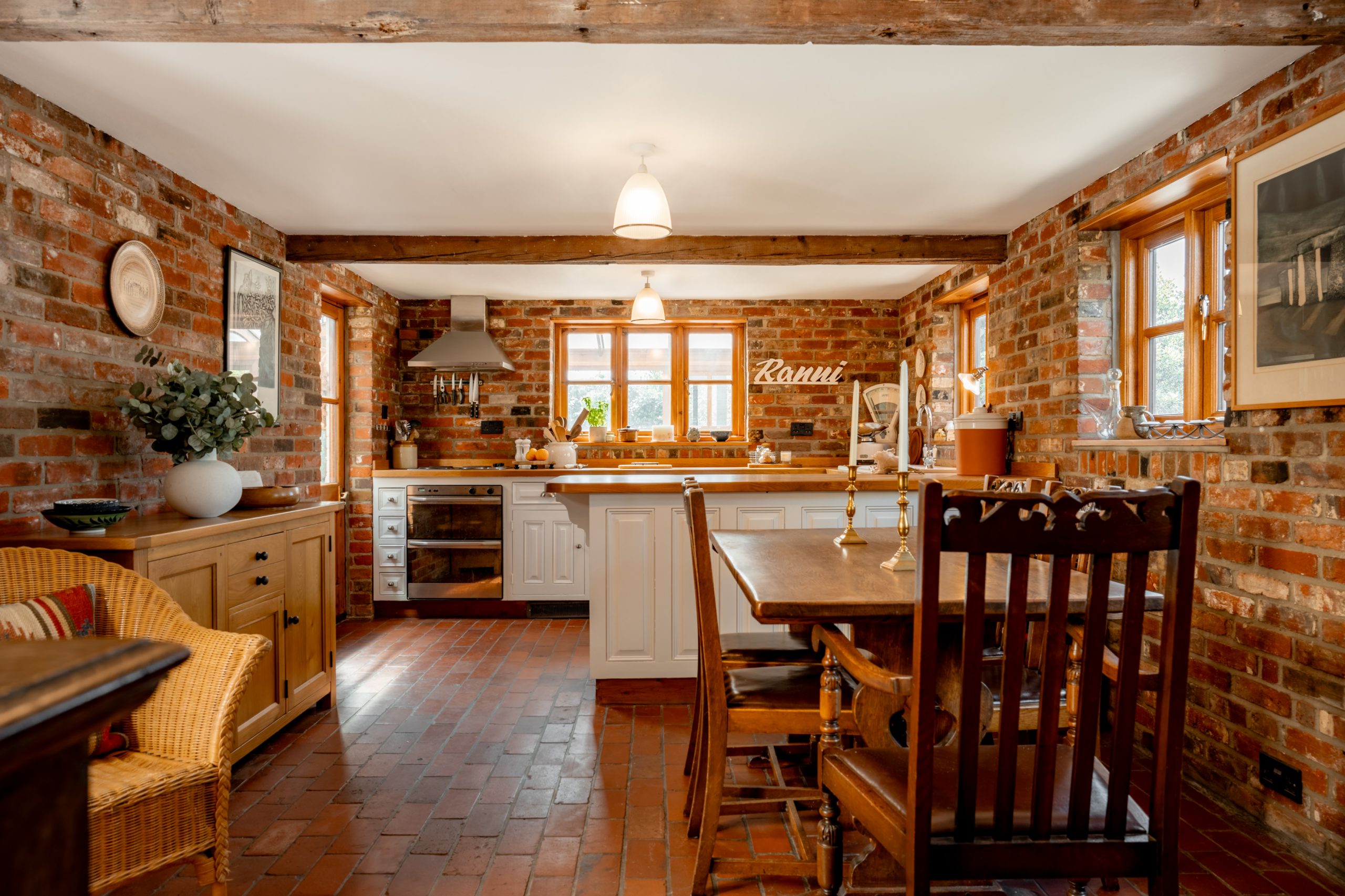 A cozy kitchen with exposed brick walls, wood beams, and rustic decor. White cabinets and modern appliances are paired with wooden furniture, including a dining table and chairs. Large windows let in natural light, highlighting the warm and inviting atmosphere.