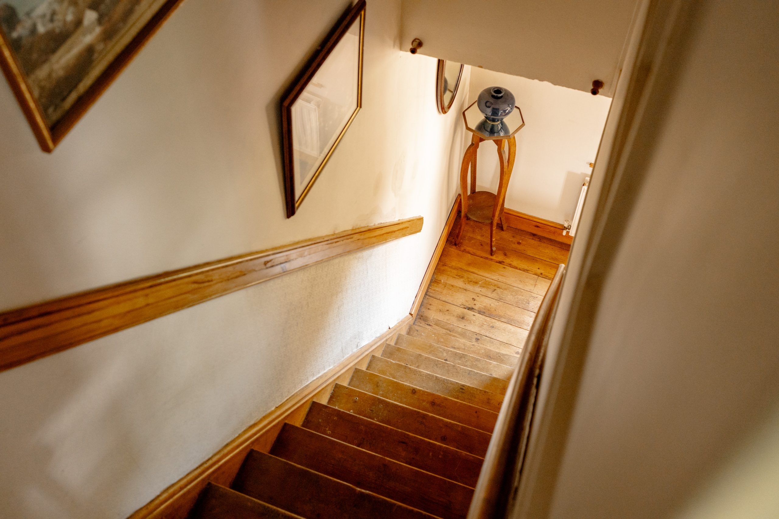 An interior view of a wooden staircase descending to a small landing area. The staircase has wooden handrails on one side. A wooden stand with a blue-and-white patterned table lamp is visible at the bottom of the stairs. The walls have framed artwork.