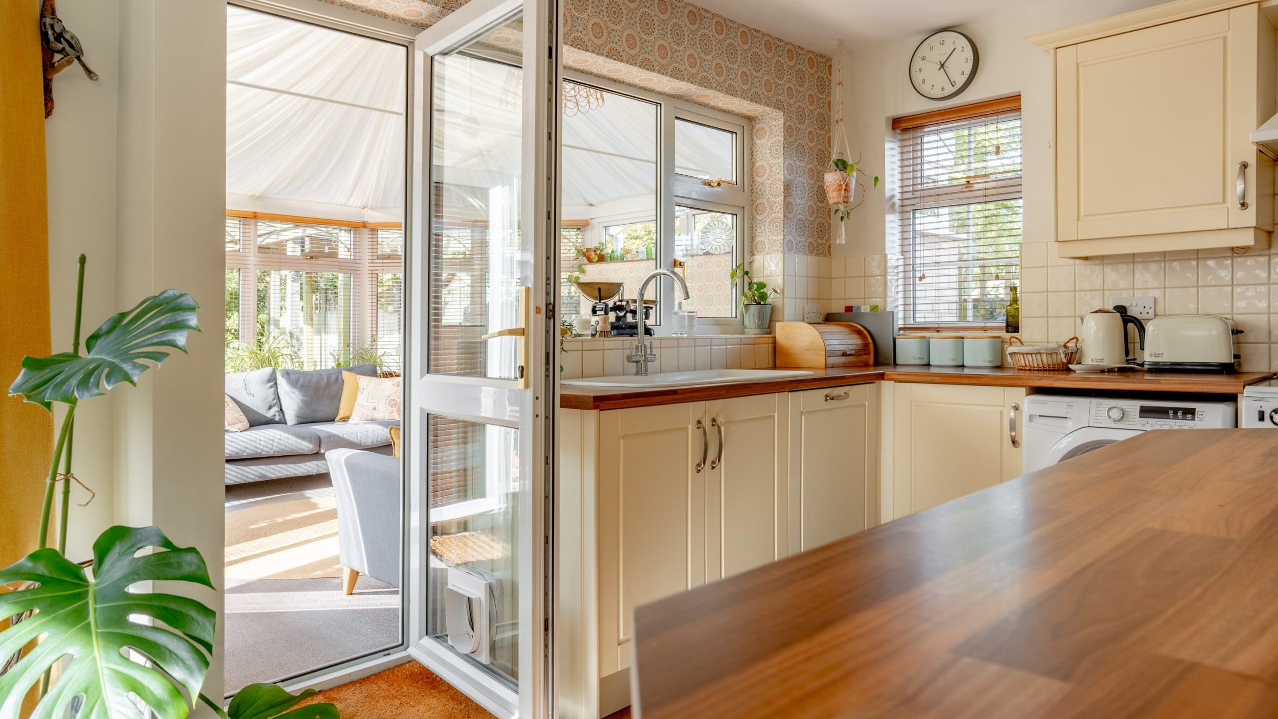 A bright kitchen featuring wooden countertops, cream cabinets, and a clock on the wall. The kitchen opens into a sunlit conservatory with seating. Near the foreground, a leafy plant adds a touch of greenery.