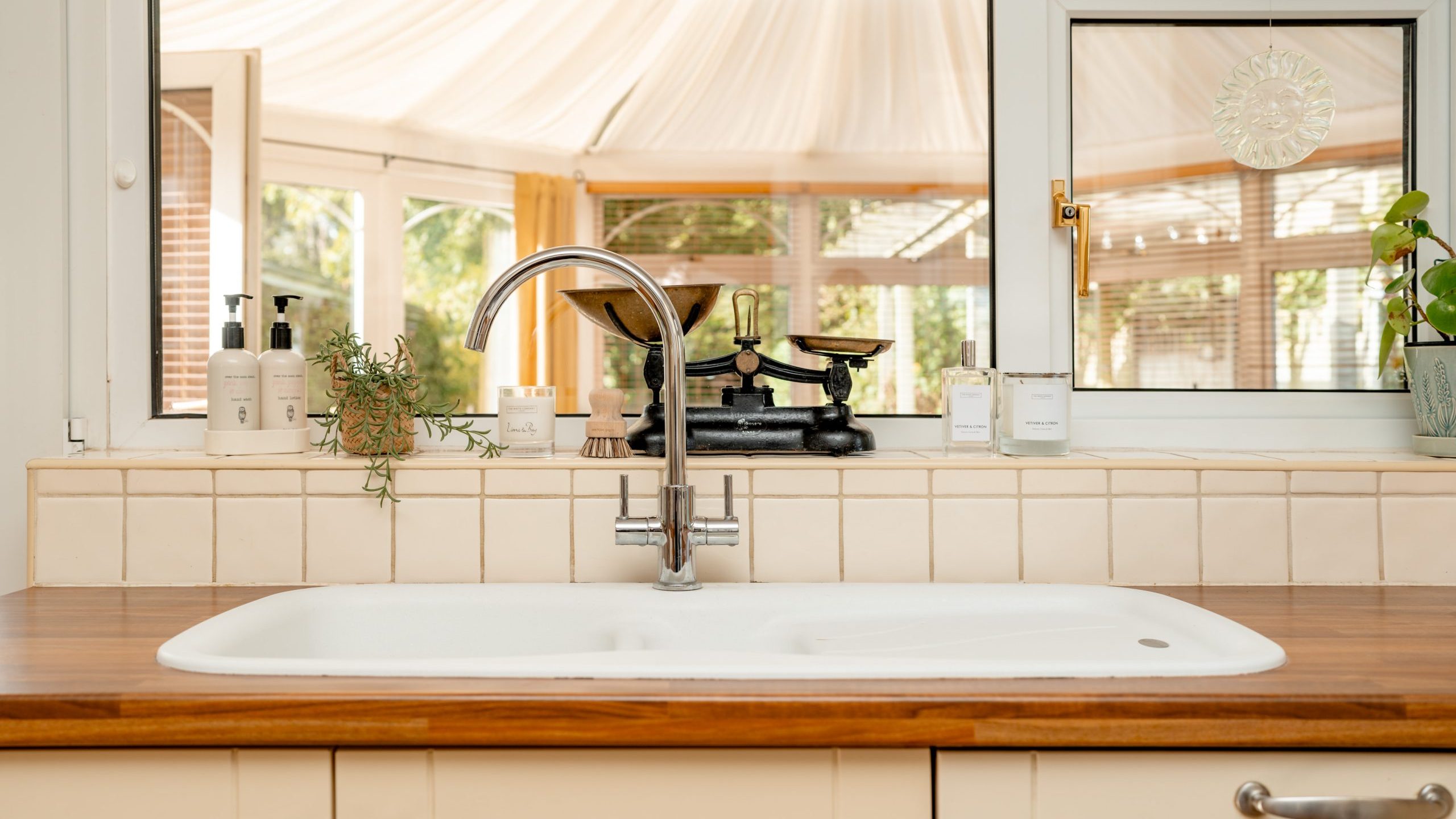 A bright kitchen sink with a wooden countertop, featuring a silver faucet, soap dispensers, a small plant, and vintage scales. In the background, a sunlit room with large windows and light curtains is visible.