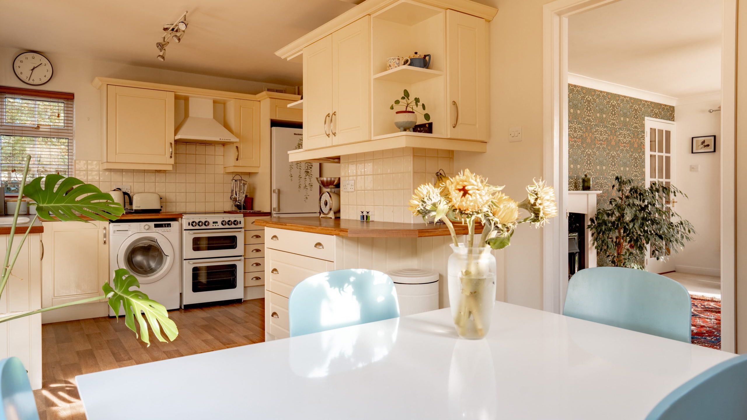 A cozy kitchen with cream cabinets, a white stove, and a washing machine. A white dining table with a vase of flowers and blue chairs is in the foreground. Sunlight filters in, illuminating the wooden floor and adjacent room.
